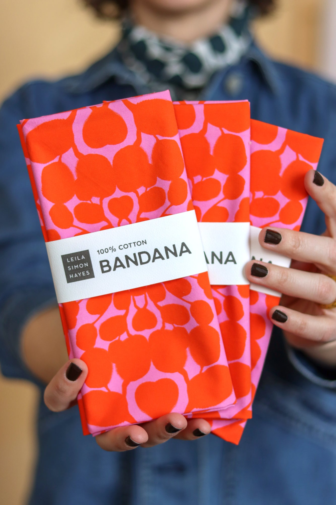Person holding three red and pink patterned bandanas with a visible brand label. Designed by artist Leila Simon Hayes.