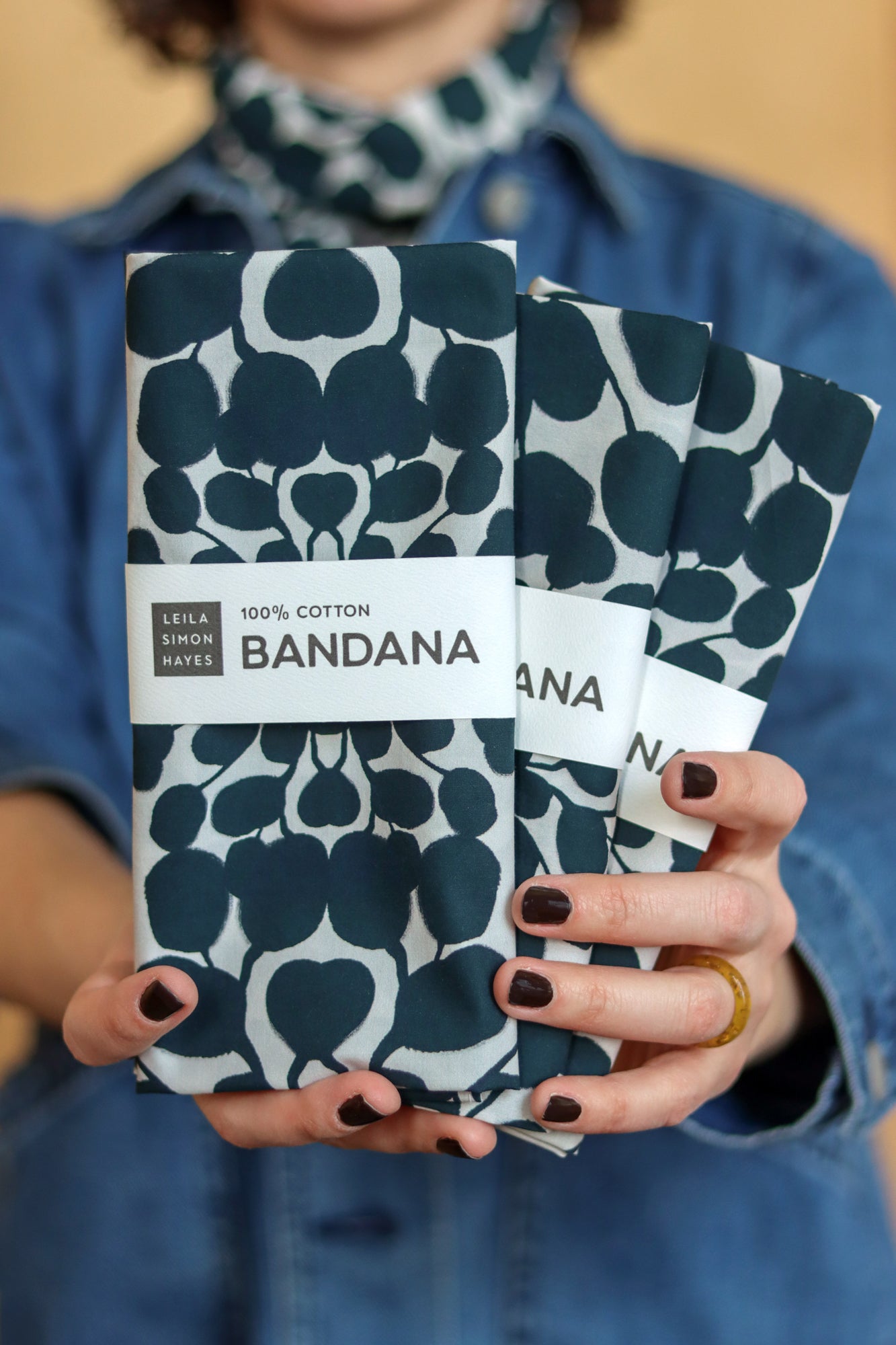 Person holding two patterned bandanas with a visible brand label. Designed by artist Leila Simon Hayes.