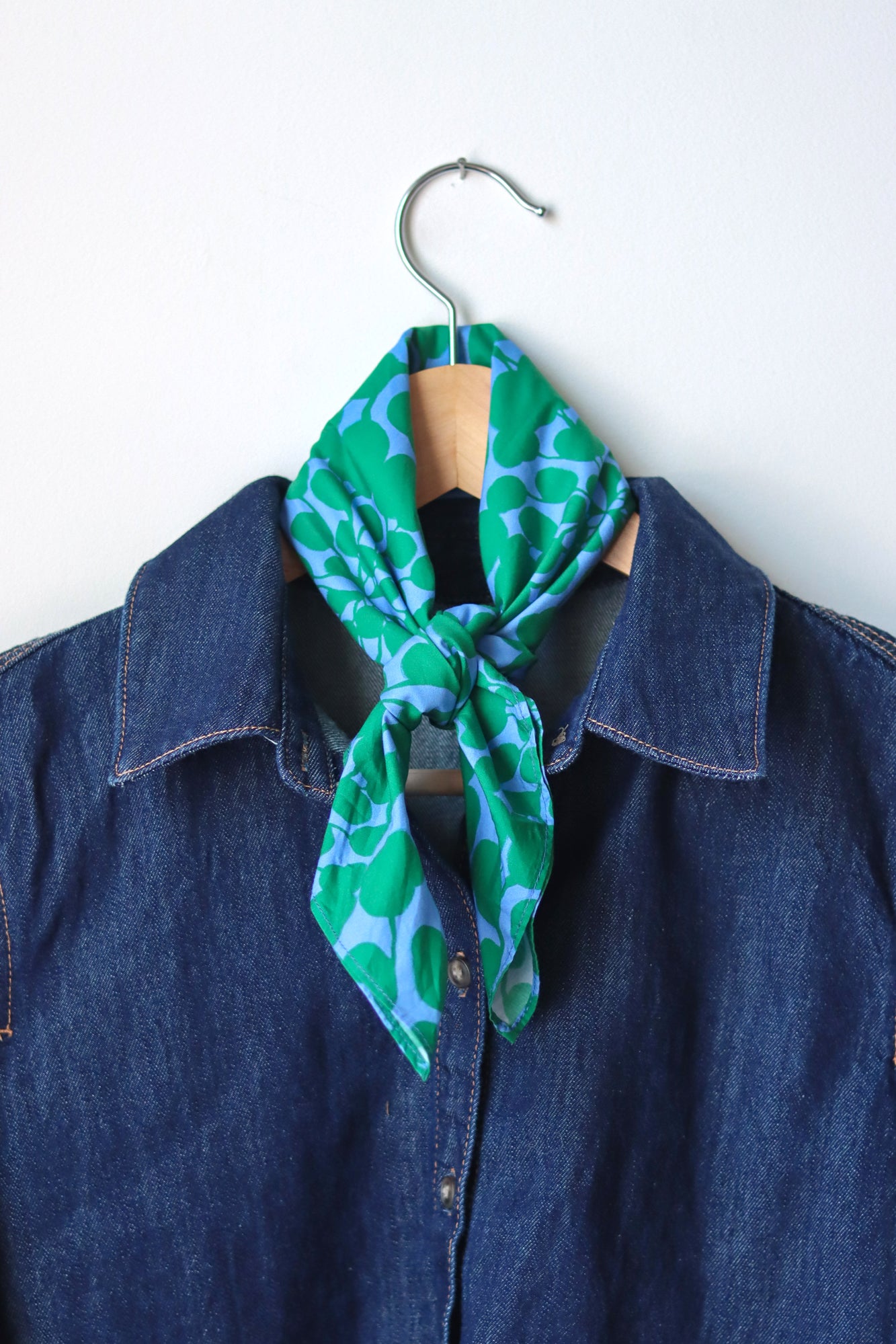 Green patterned bandana tied into a scarf draped over a blue denim shirt on a hanger against a white background. Designed by artist Leila Simon Hayes.