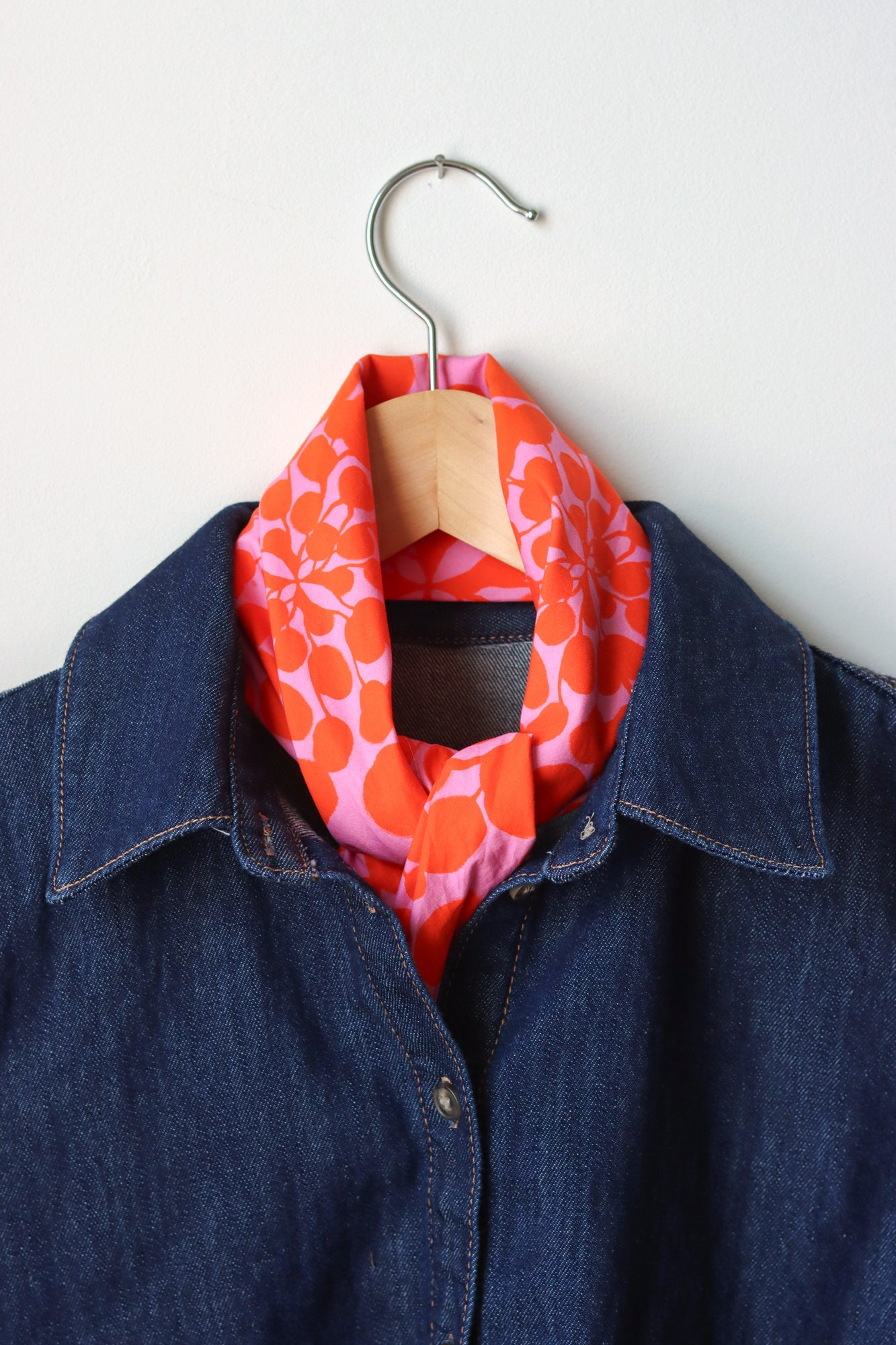 Orange and pink patterned bandana tied into a scarf draped over a denim jacket on a hanger against a white background. Designed by artist Leila Simon Hayes.