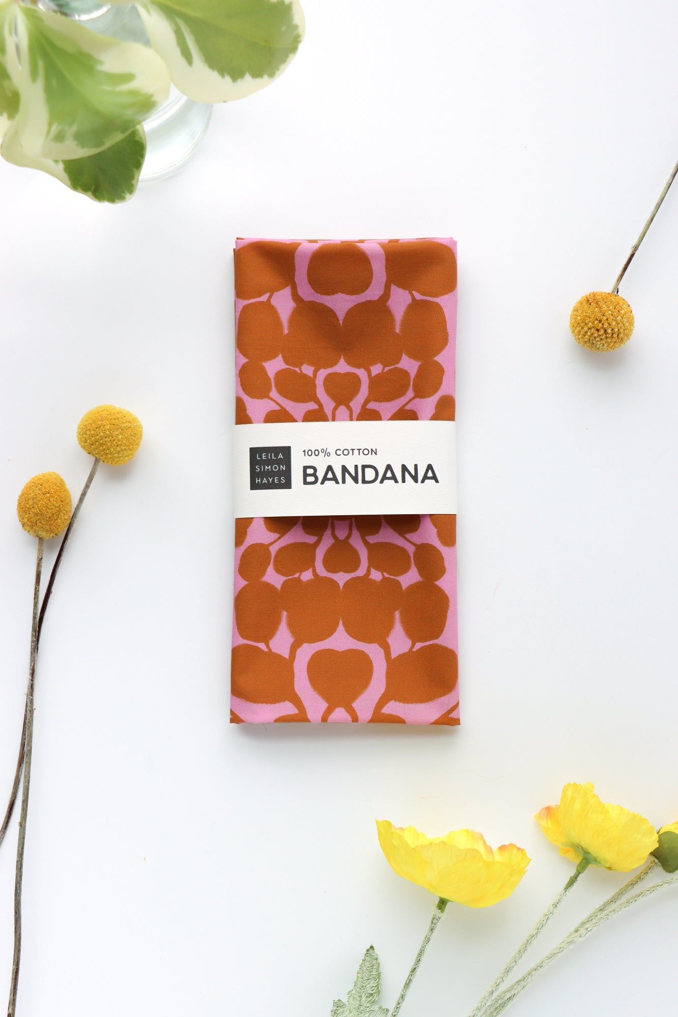 Packaged bandana with pink and brown pattern on a white background with flowers. Designed by artist Leila Simon Hayes.