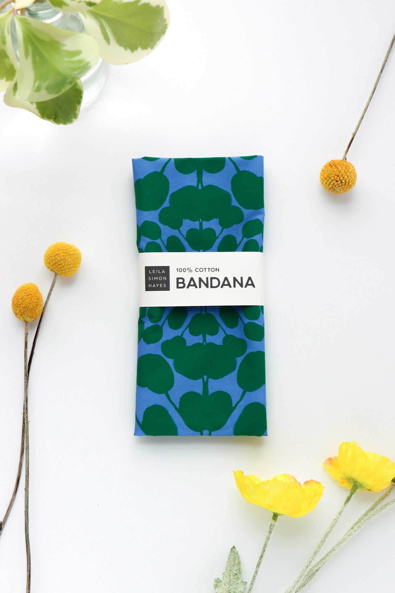 Blue and green patterned bandana with a white label on a white background with yellow flowers and green leaves. Designed by artist Leila Simon Hayes.