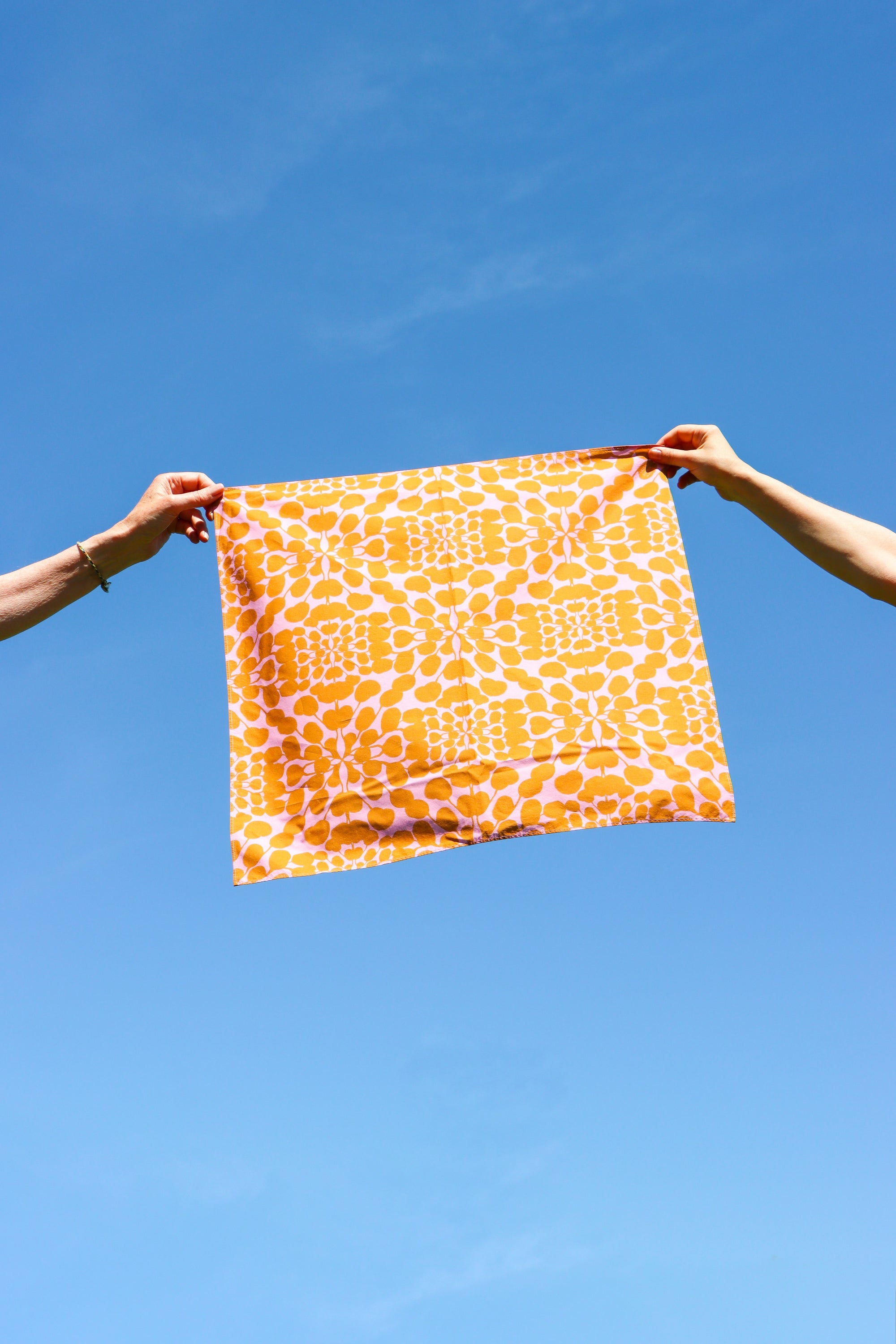 An orange and white patterned square scarf is held up by two hands against a clear blue sky. Designed by artist Leila Simon Hayes.