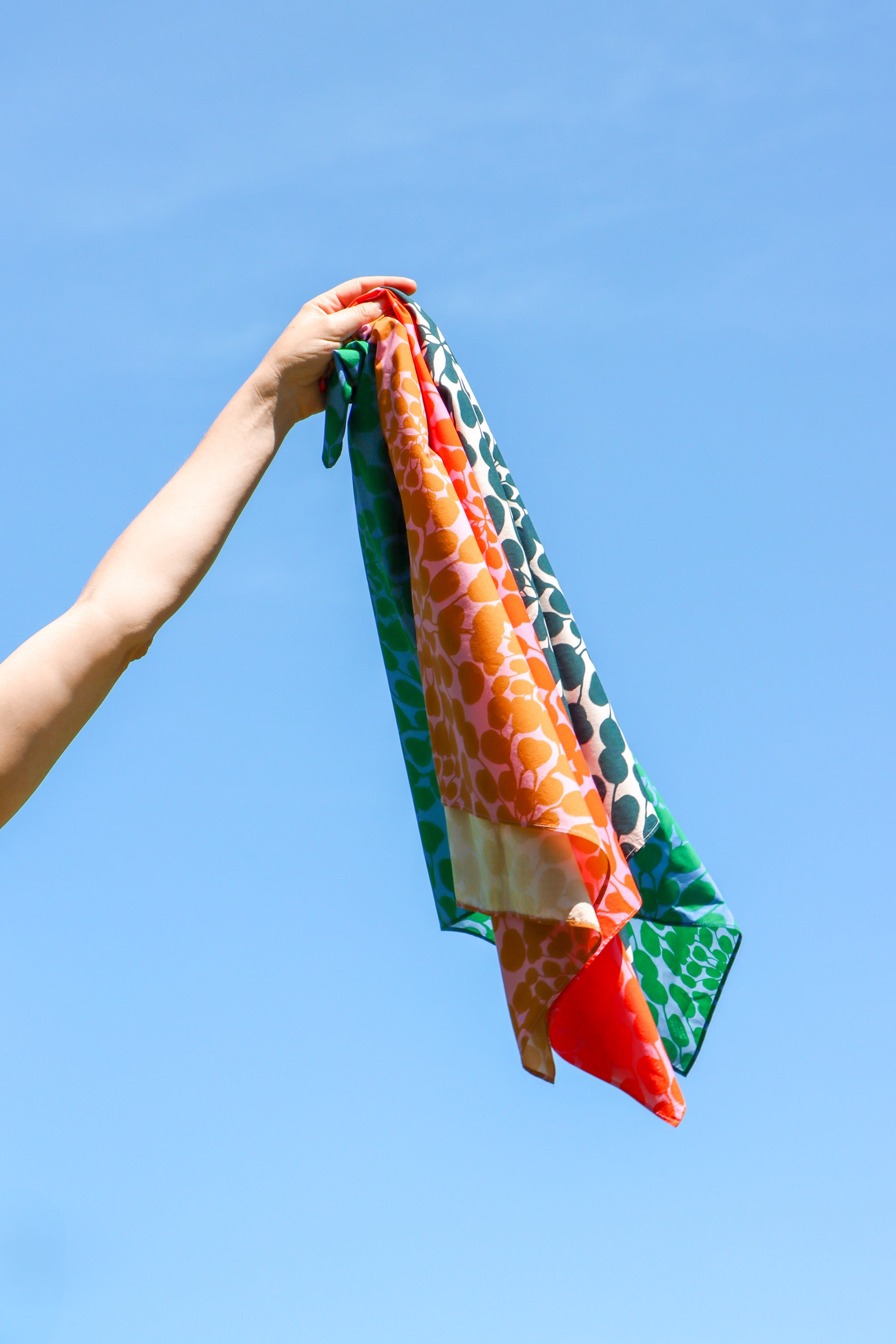 A hand holding a lightweight scarf against a clear blue sky. The scarf features a multicolor pattern with orange, green, red, and cream sections. Designed by artist Leila Simon Hayes.