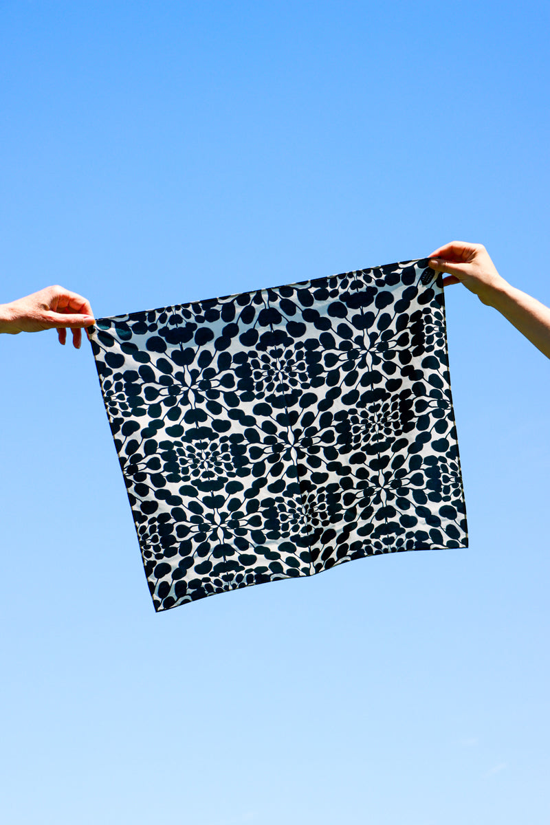Person holding a black and white bandana with floral print fabric against a clear blue sky. Designed by artist Leila Simon Hayes.