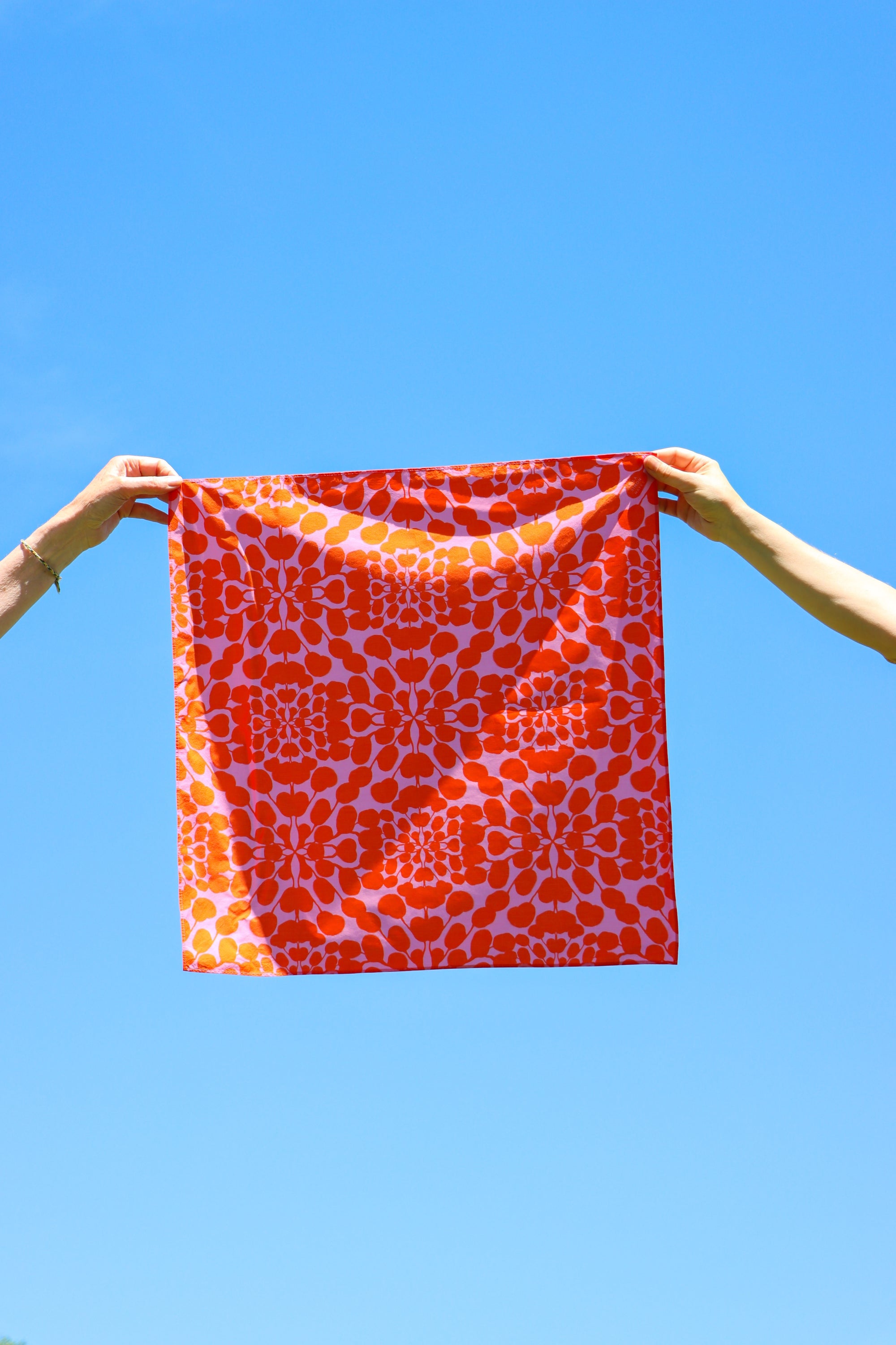A square bandana with a vibrant orange and white geometric pattern is held up by two hands against a clear blue sky. Designed by artist Leila Simon Hayes.