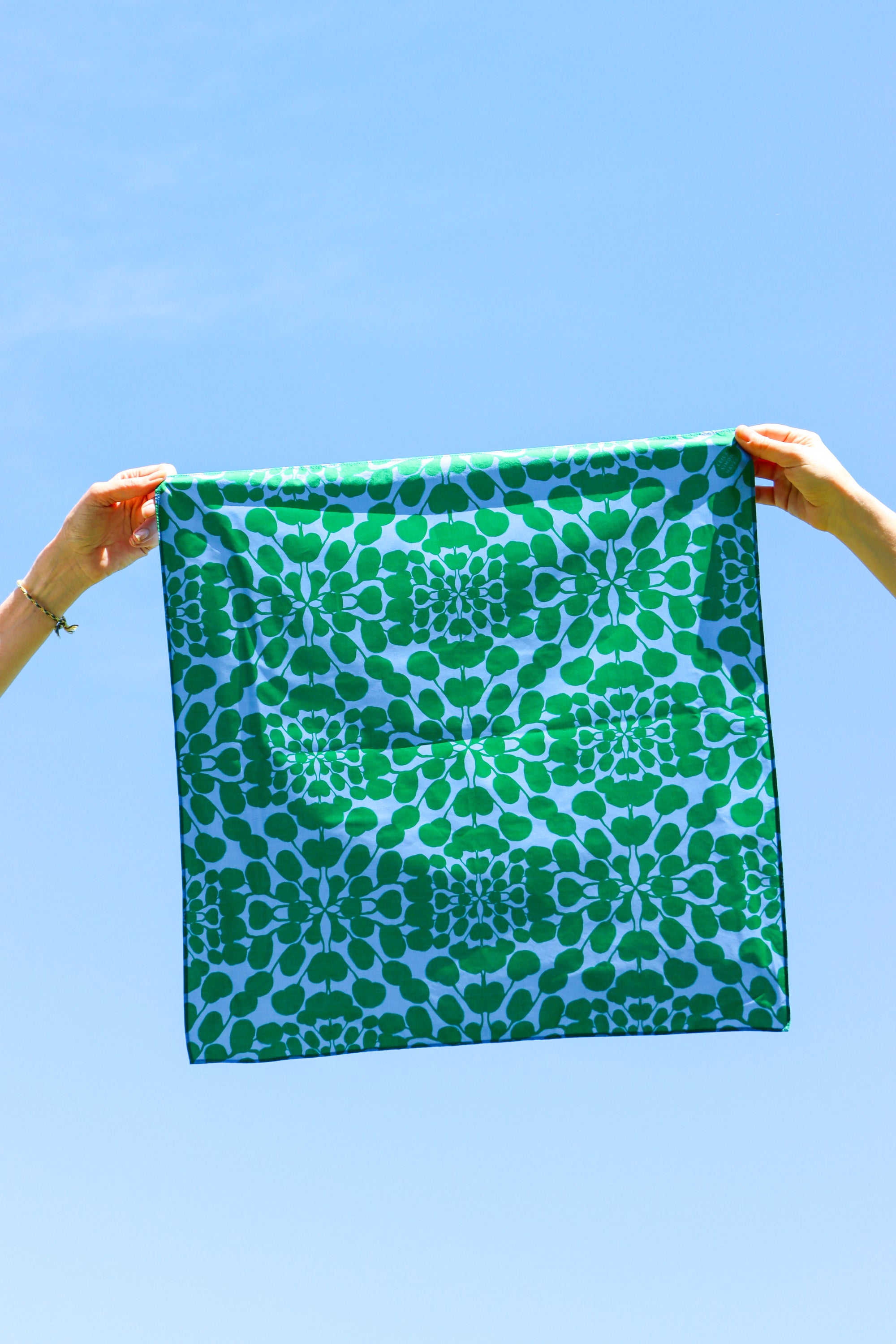 A square scarf with a green and light blue geometric floral pattern is held up by two hands against a clear blue sky. Designed by artist Leila Simon Hayes.