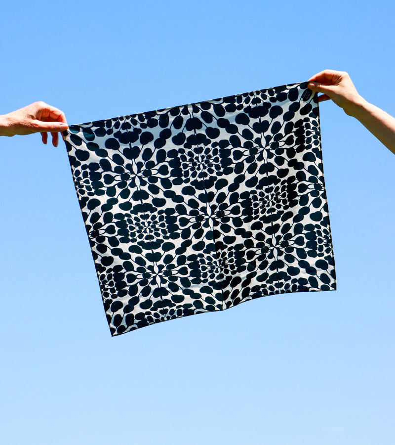 A square, cotton bandana designed by Leila Simon Hayes with a black and white abstract organic pattern, held by two hands against a clear blue sky.
