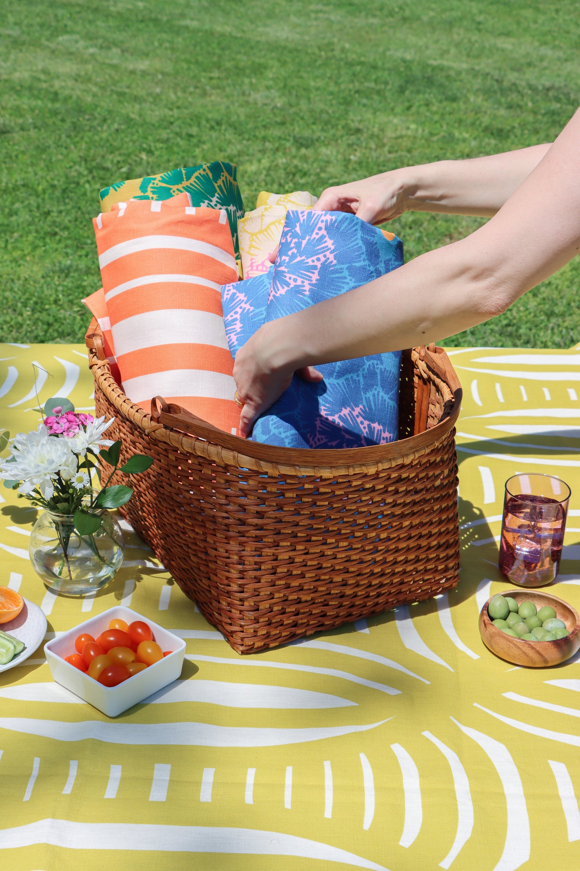 A wicker picnic basket filled with colorful patterned towels or blankets, set on a yellow-and-white picnic blanket outdoors on grass. A person's arms are visible reaching into the basket, with small food items and flowers nearby. Designed by artist Leila Simon Hayes.