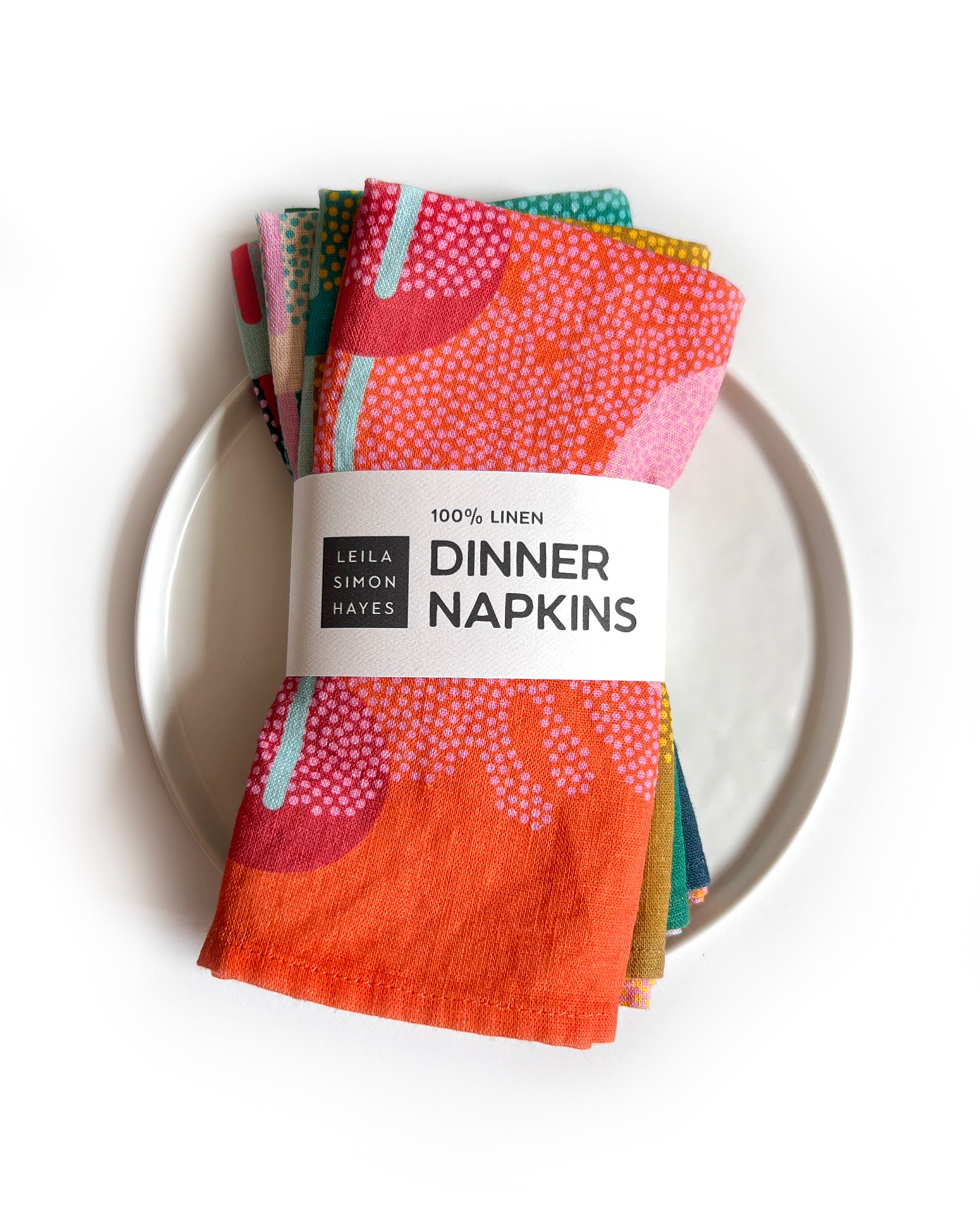 Colorful pack of four dinner napkins on a white plate with a Leila Simon Hayes label saying "100% Linen Dinner Napkins".