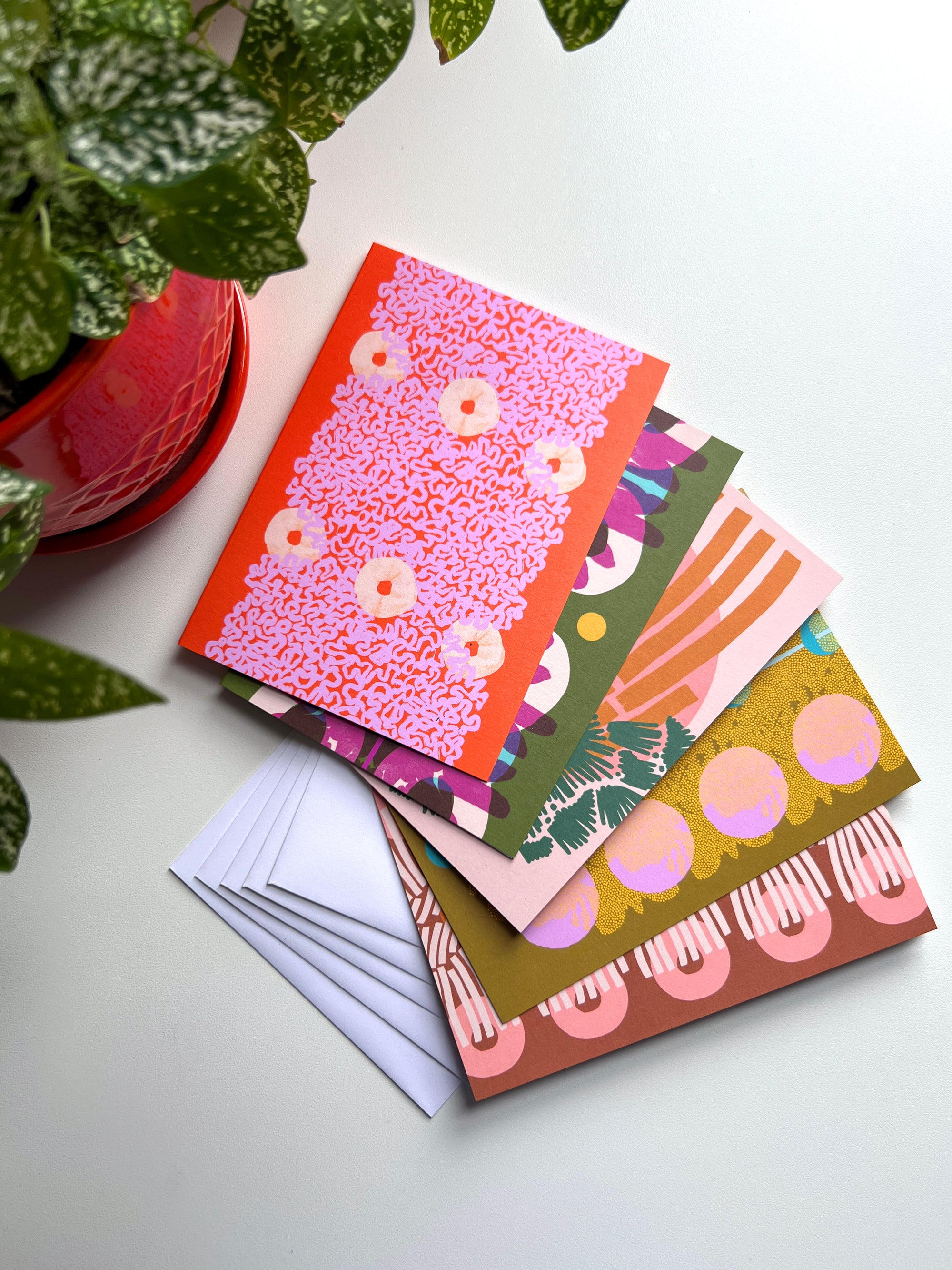 A set of five folded note cards with various colorful abstract art designs, displayed with envelopes on a table, with a red potted plant in the background. Designed by artist Leila Simon Hayes.