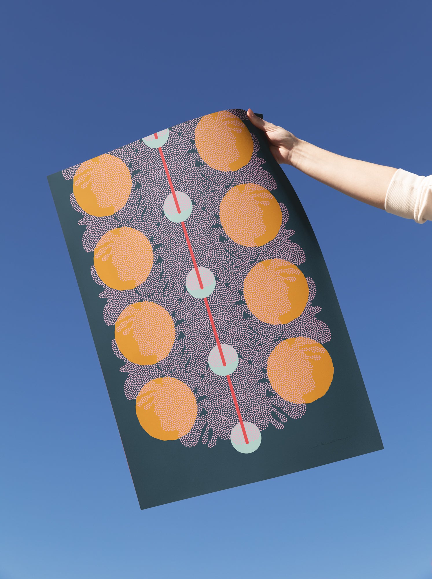 A hand holds up a large art print against a clear blue sky. The print features a dark teal background with a vertical pattern of orange circles, light blue dots, and a red line.