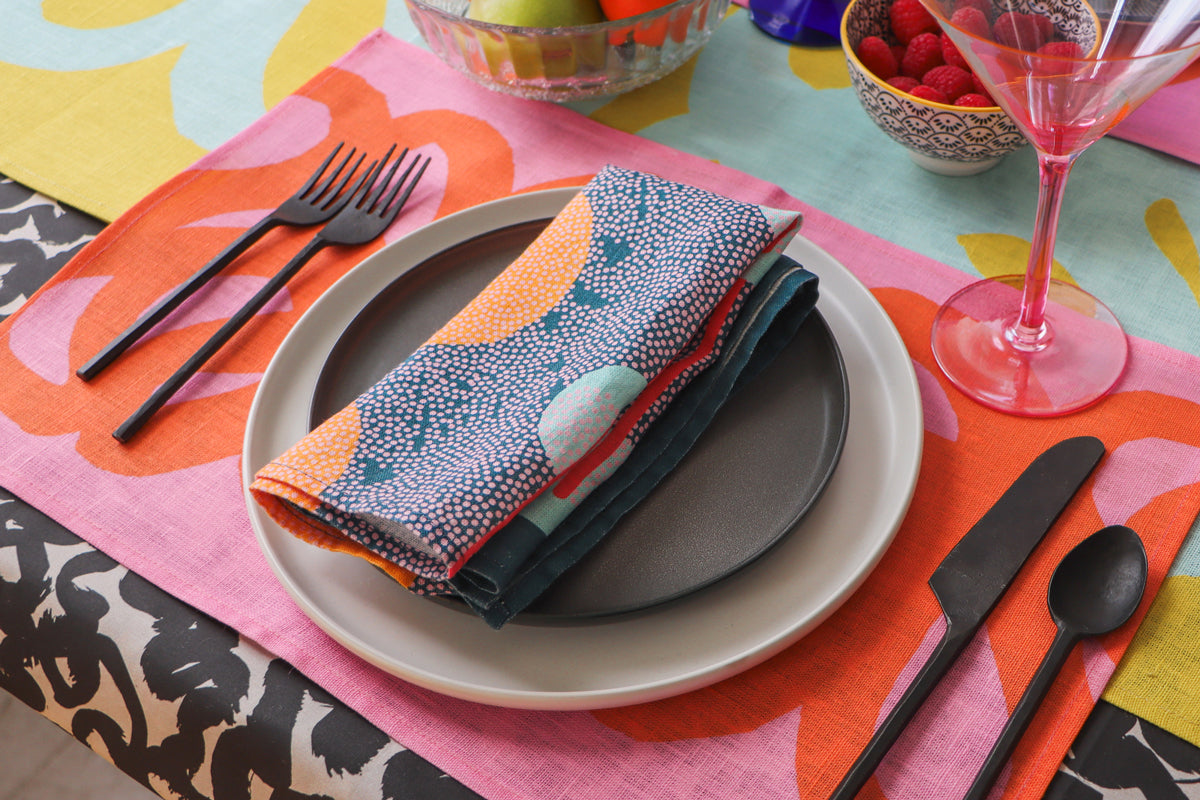 A set table with a blue linen napkin placed on a plate, surrounded by colorful table linens including a pink and coral placemat, floral table runner, and an abstract tablecloth. Designed by artist Leila Simon Hayes. 