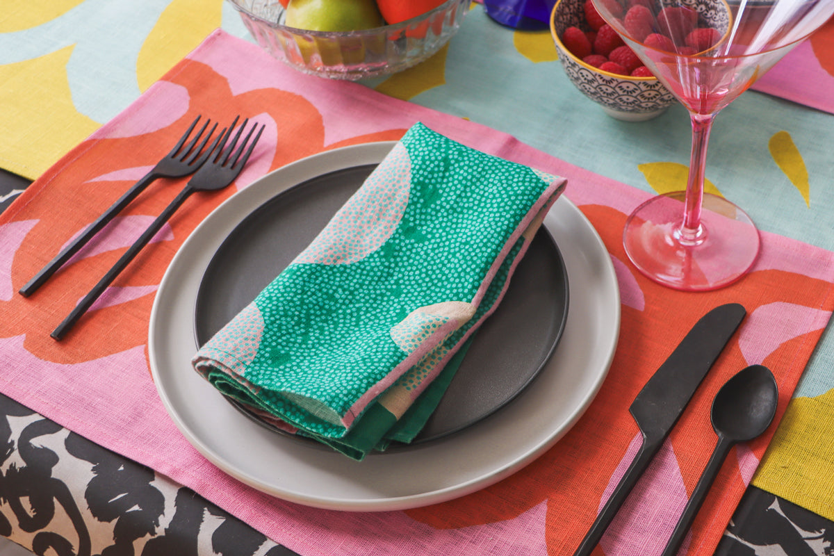 A green linen napkin is displayed on a plate, with a colorful patterned placemat, table runner and tablecloth underneath. Designed by artist Leila Simon Hayes. 