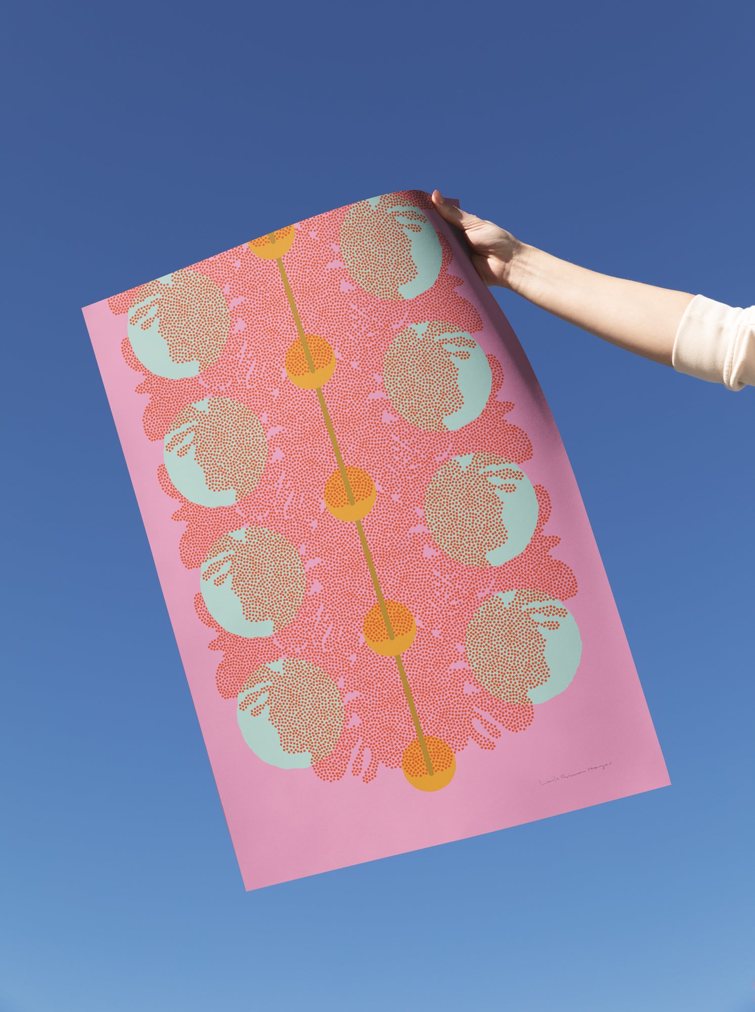 A hand holds a large art print against a clear blue sky. The print features a pink background with a central vertical gold line, repeating circular motifs in pale mint green and coral, and small gold dots along the line.
