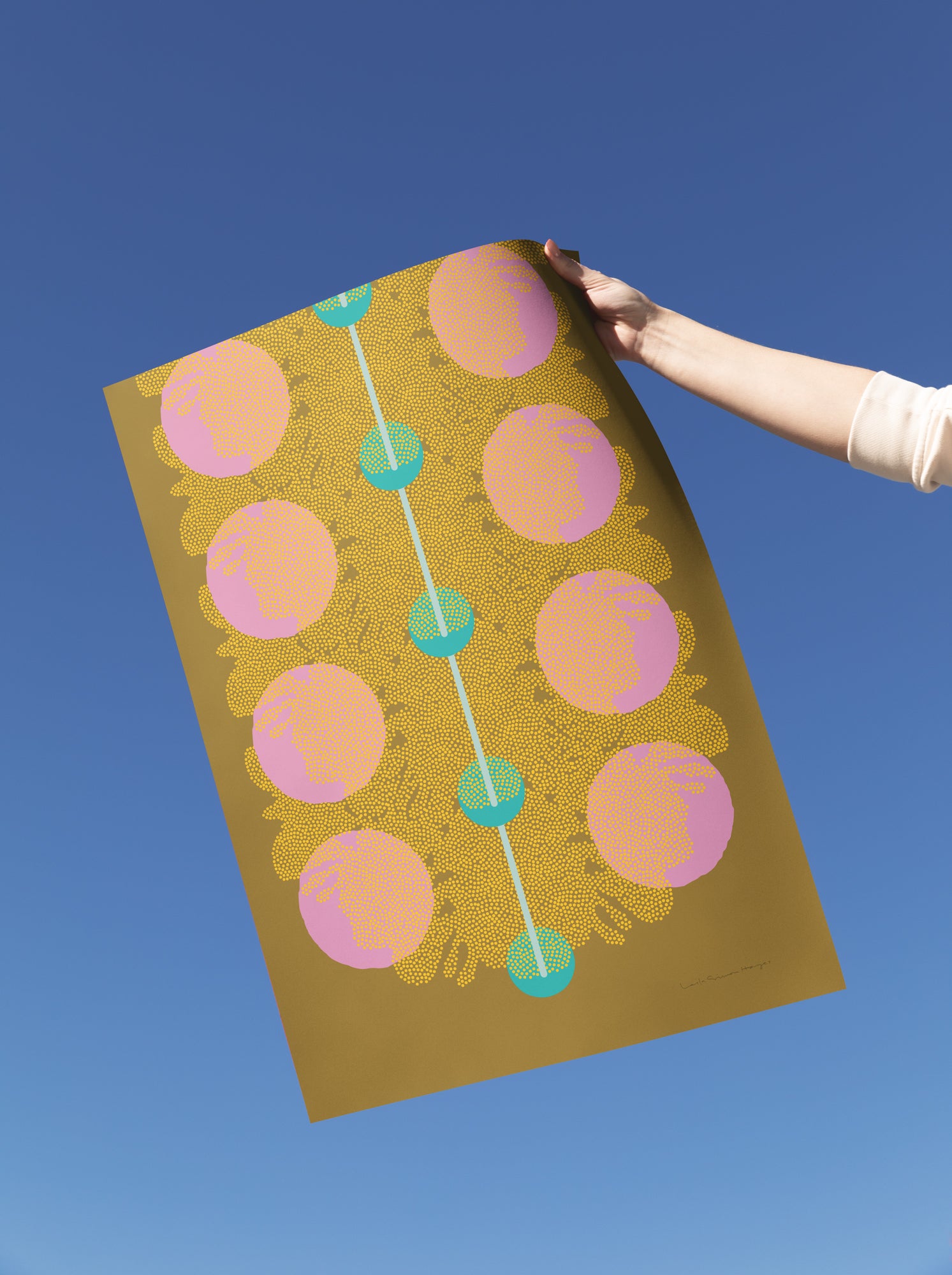 A hand holds a large art print against a clear blue sky. The print features a gold background with a vertical line of pink circles connected by teal dots. Designed by artist Leila Simon Hayes.