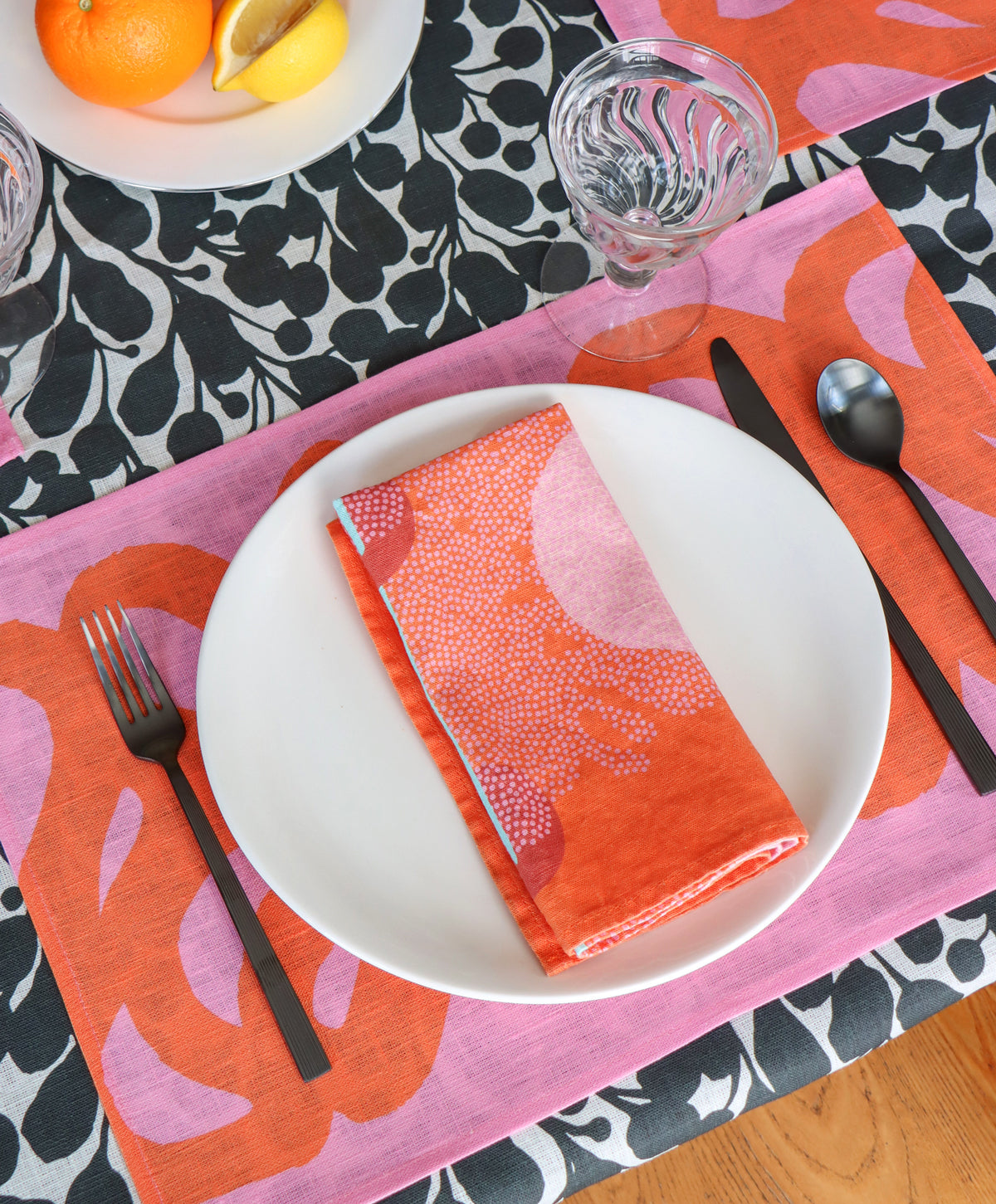 A coral patterned linen napkin is displayed on a white plate, with a pink and coral patterned linen placemat and black and white botanical tablecloth underneath. 