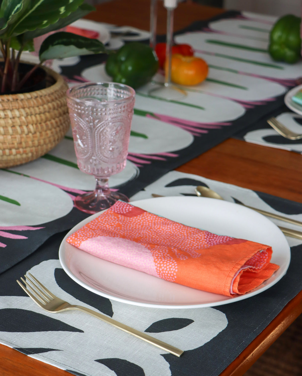A table set with table linens. A red napkin on a plate over a black and white placemat and an abstract black, white, pink and green table runner. A lifestyle image of what your table could look like with the linens designed by artist Leila Simon Hayes