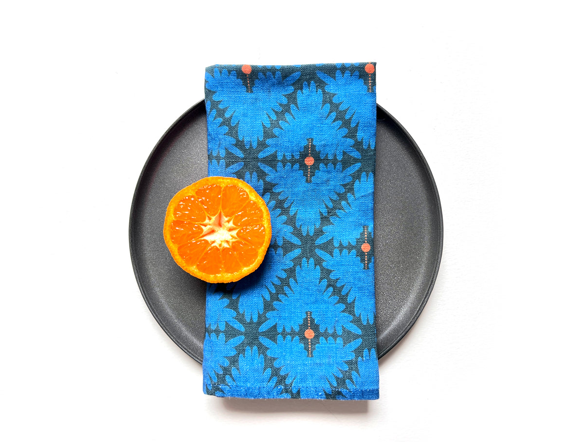 A folded blue patterned cloth napkin with a geometric ikat design and small coral accents, displayed on a black plate next to a halved orange on a white background. Designed by artist Leila Simon Hayes. 