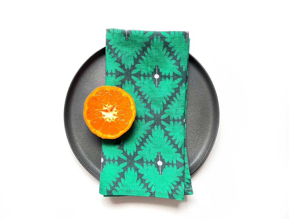 A folded green napkin with a dark gray geometric ikat pattern and small white dot accents, styled on a black plate next to a halved orange on a white background. Designed by artist Leila Simon Hayes. 
