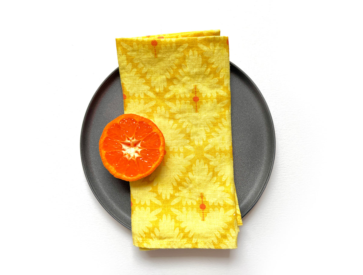 A folded yellow patterned cloth napkin with a small orange slice on top, arranged on a round dark gray plate against a white background. Designed by artist Leila Simon Hayes. 