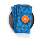 A stack of blue patterned cloth napkins with a dark geometric print and small orange dots, arranged on a black plate with a halved orange on top, photographed on a white background. Designed by artist Leila Simon Hayes.