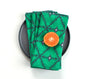 A stack of green napkins with a dark gray geometric pattern, styled on a black plate with a sliced orange on a white background. Designed by artist Leila Simon Hayes.