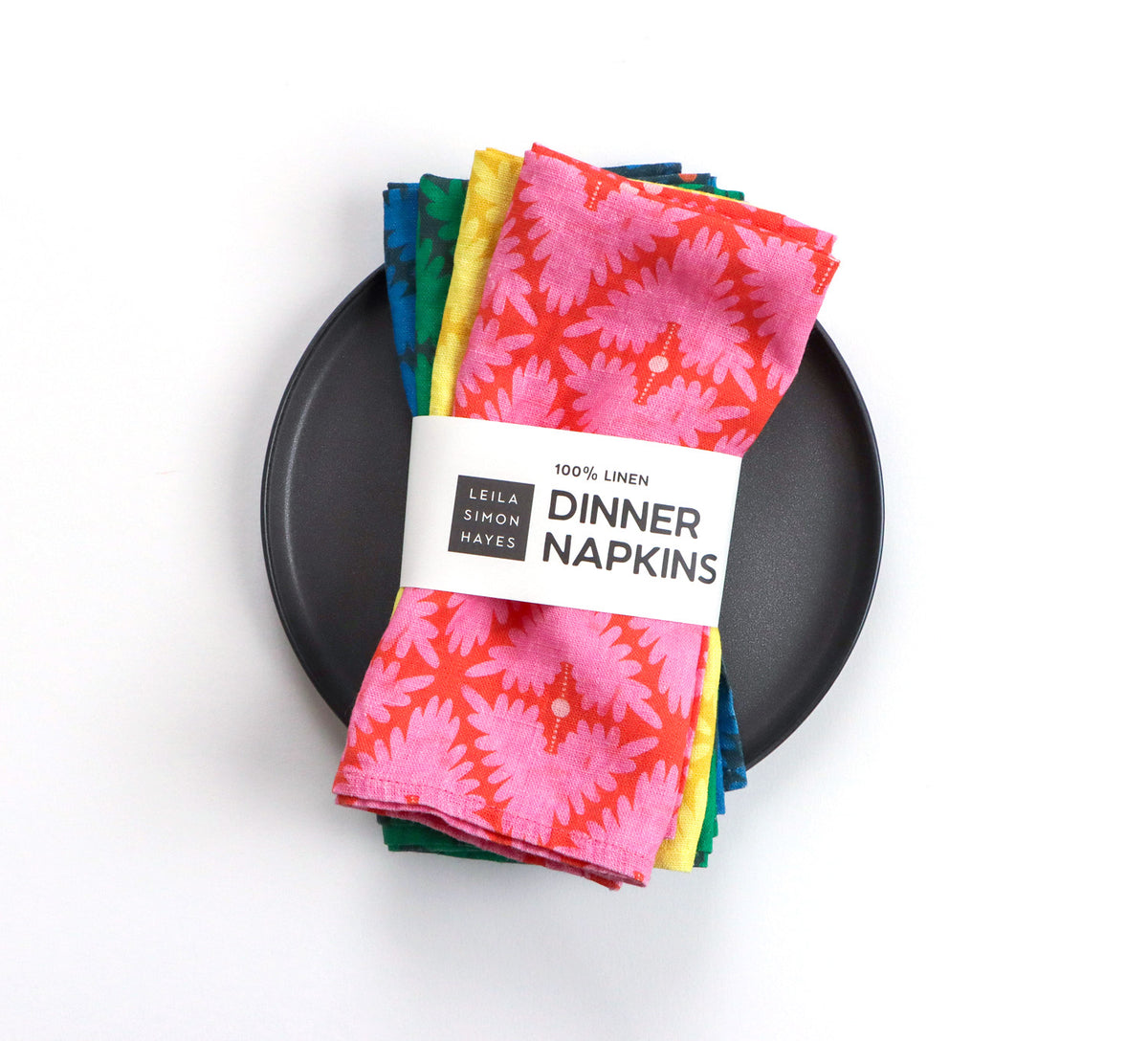 A stack of colorful patterned linen napkins wrapped in a white paper band that reads '100% LINEN LEILA SIMON HAYES DINNER NAPKINS,' placed on a round black plate against a clean white background. Designed by artist Leila Simon Hayes. 