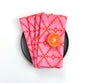 A stack of folded pink and red patterned cloth napkins on a black plate, with a halved orange placed on top, photographed from above on a white background. Designed by artist Leila Simon Hayes. 