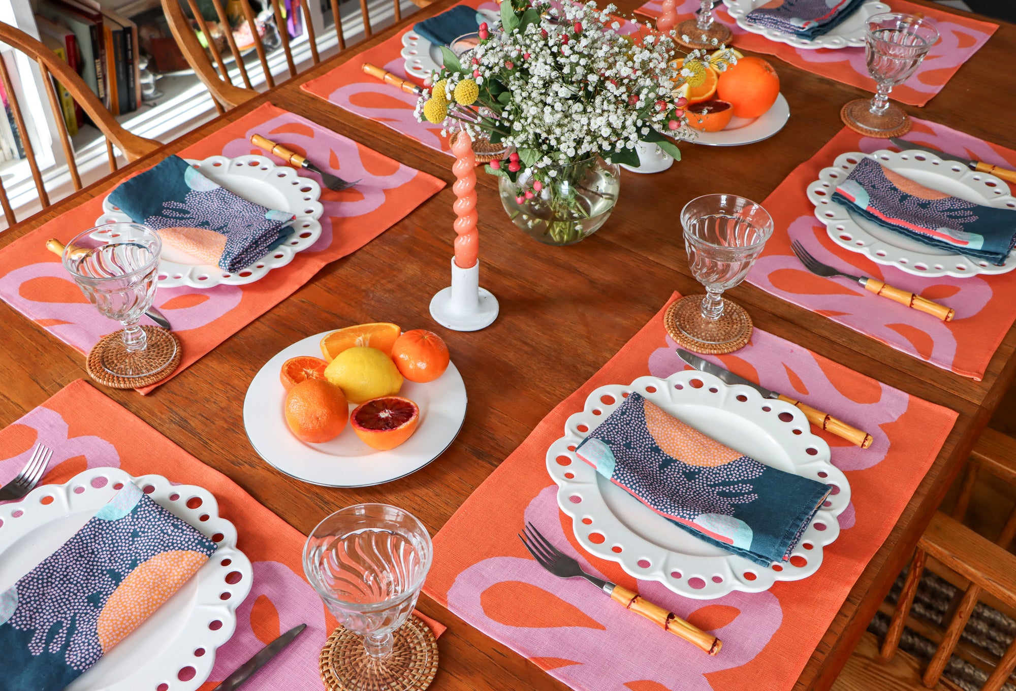 A dining room table with blue napkins, coral and pink placemats and flower, plates and cutlery. Table linens designed by artist Leila Simon Hayes