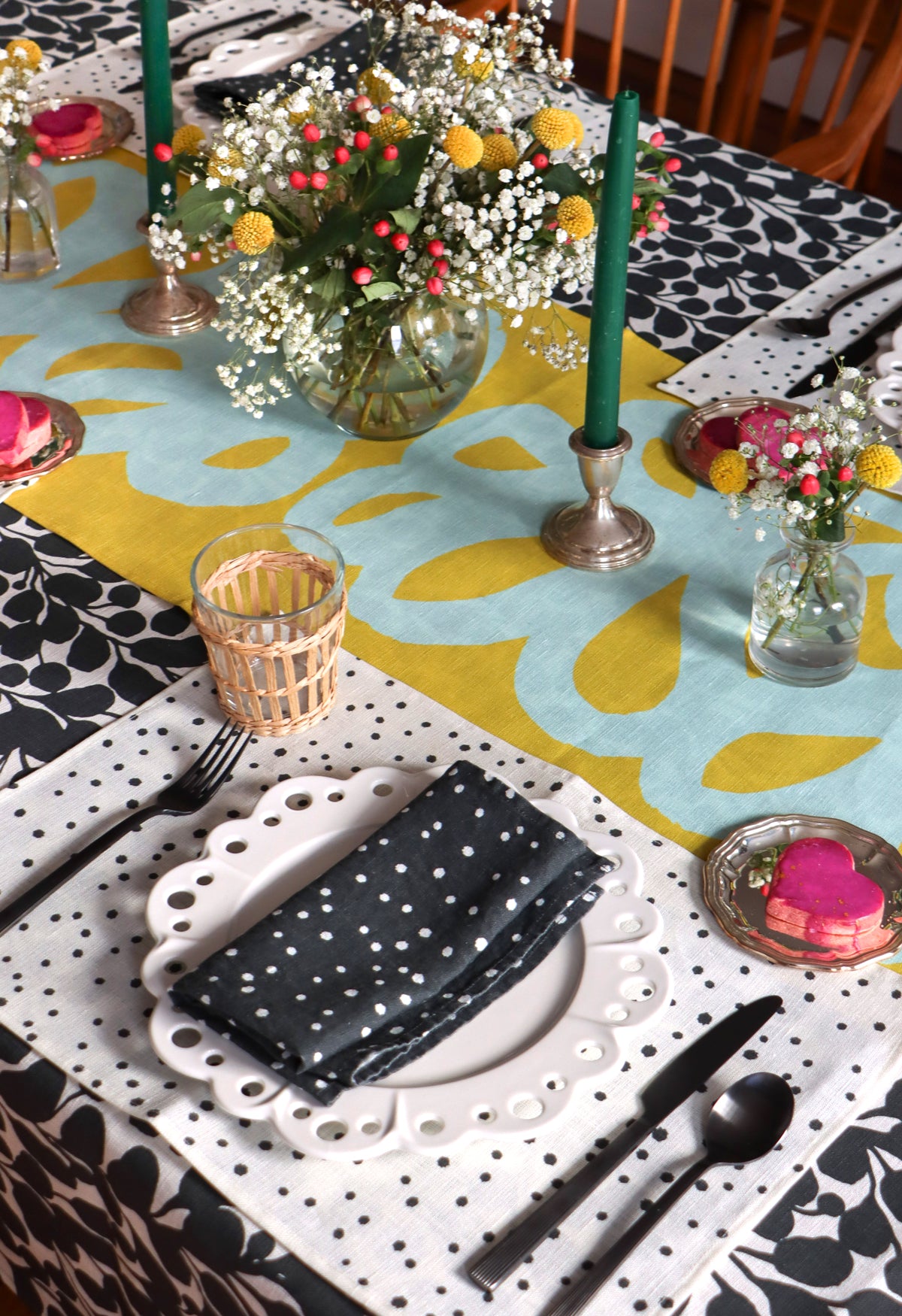 A black and white napkin, white linen placemat with a black organic dotted pattern, and green table runner displayed on a dining room table. Table linens designed by artist Leila Simon Hayes