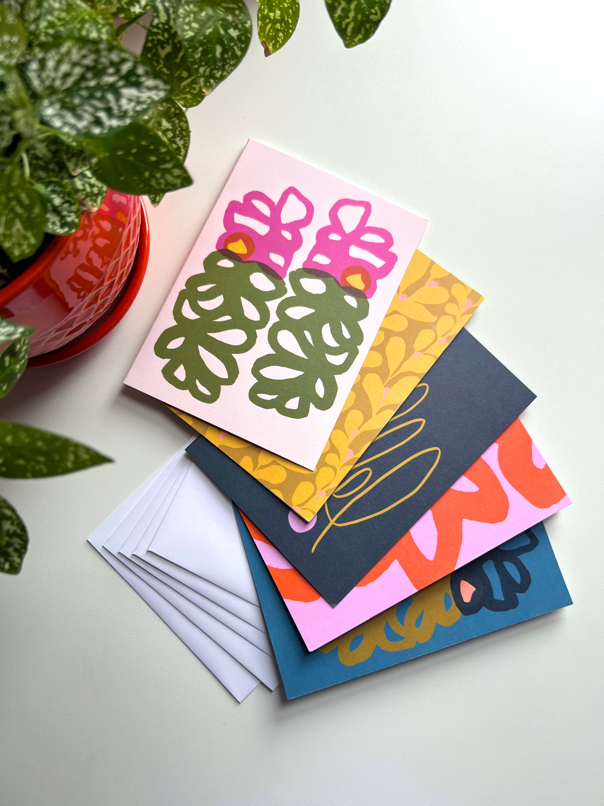 A set of five folded note cards with floral designs, displayed with envelopes and a potted plant in the background. Designed by artist Leila Simon Hayes.