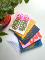 A set of five folded note cards with floral designs, displayed with envelopes and a potted plant in the background. Designed by artist Leila Simon Hayes.
