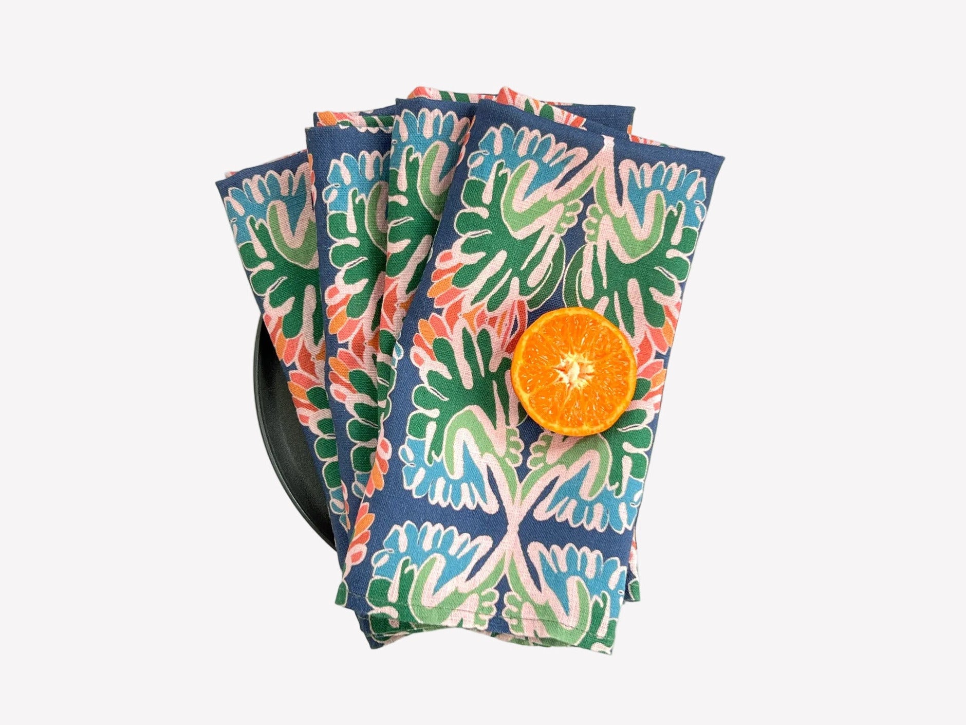 Set of colorful patterned napkins with a sliced orange on a white background. The napkin design is a floral pattern with tones of blue, green and pink. Designed by artist Leila Simon Hayes. 