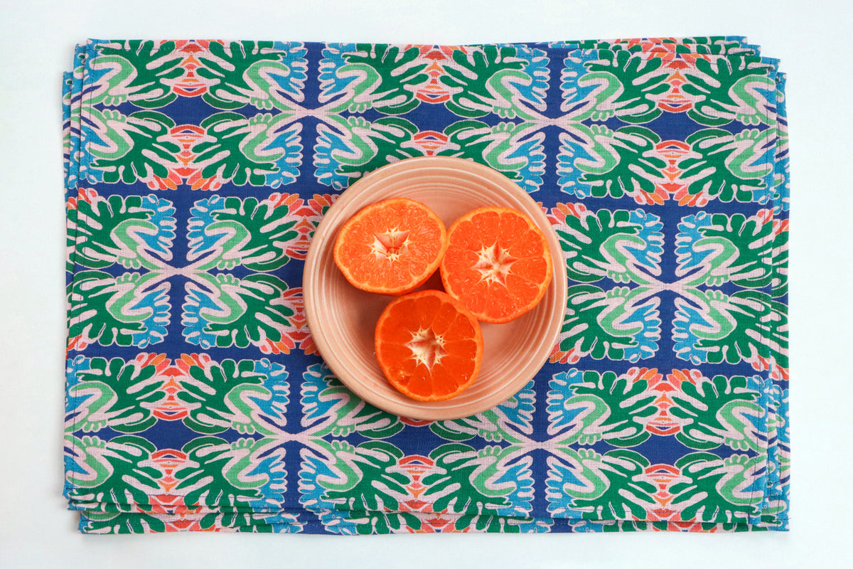 A square woven placemat with a colorful geometric tile pattern in blue, green, and coral, styled with a small beige plate holding three orange halves on a white background. Designed by artist Leila Simon Hayes.