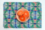 A square woven placemat with a colorful geometric tile pattern in blue, green, and coral, styled with a small beige plate holding three orange halves on a white background. Designed by artist Leila Simon Hayes.