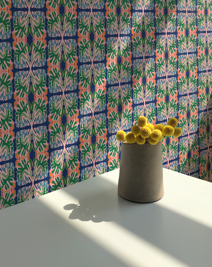 A gray ceramic vase filled with small yellow pom-pom flowers sits on a white table in front of a colorful patterned wallpaper featuring blue, green, and coral floral motifs. Designed by artist Leila Simon Hayes.