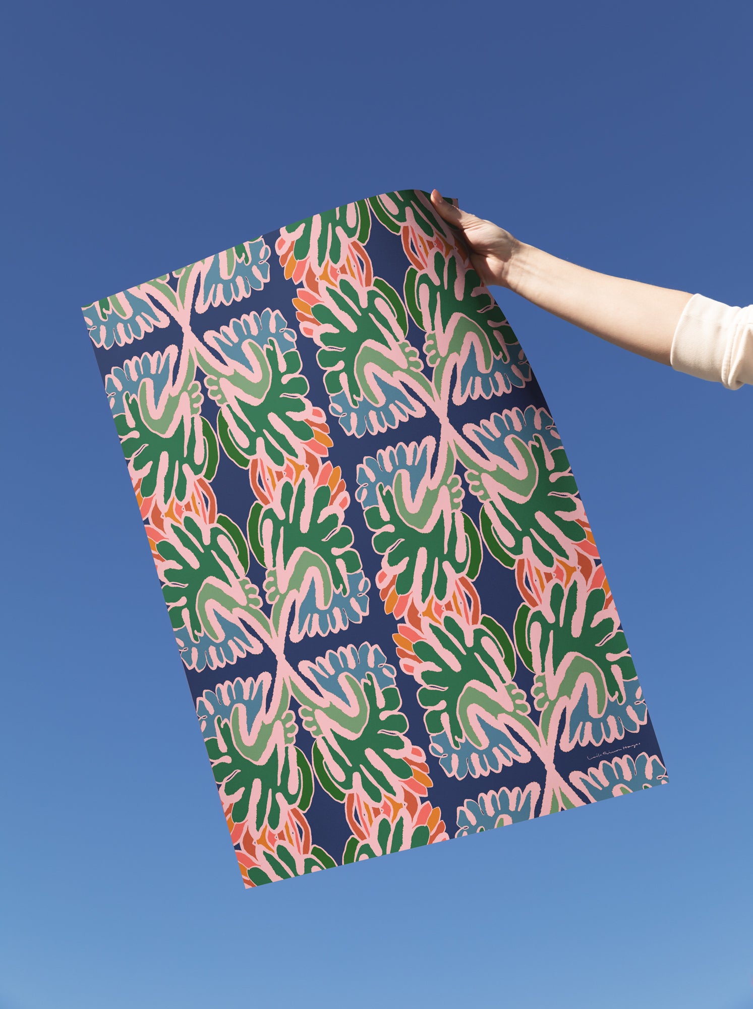 A person's arm holding up a fine art print with a colorful abstract botanical pattern against a clear blue sky. Designed by artist Leila Simon Hayes.