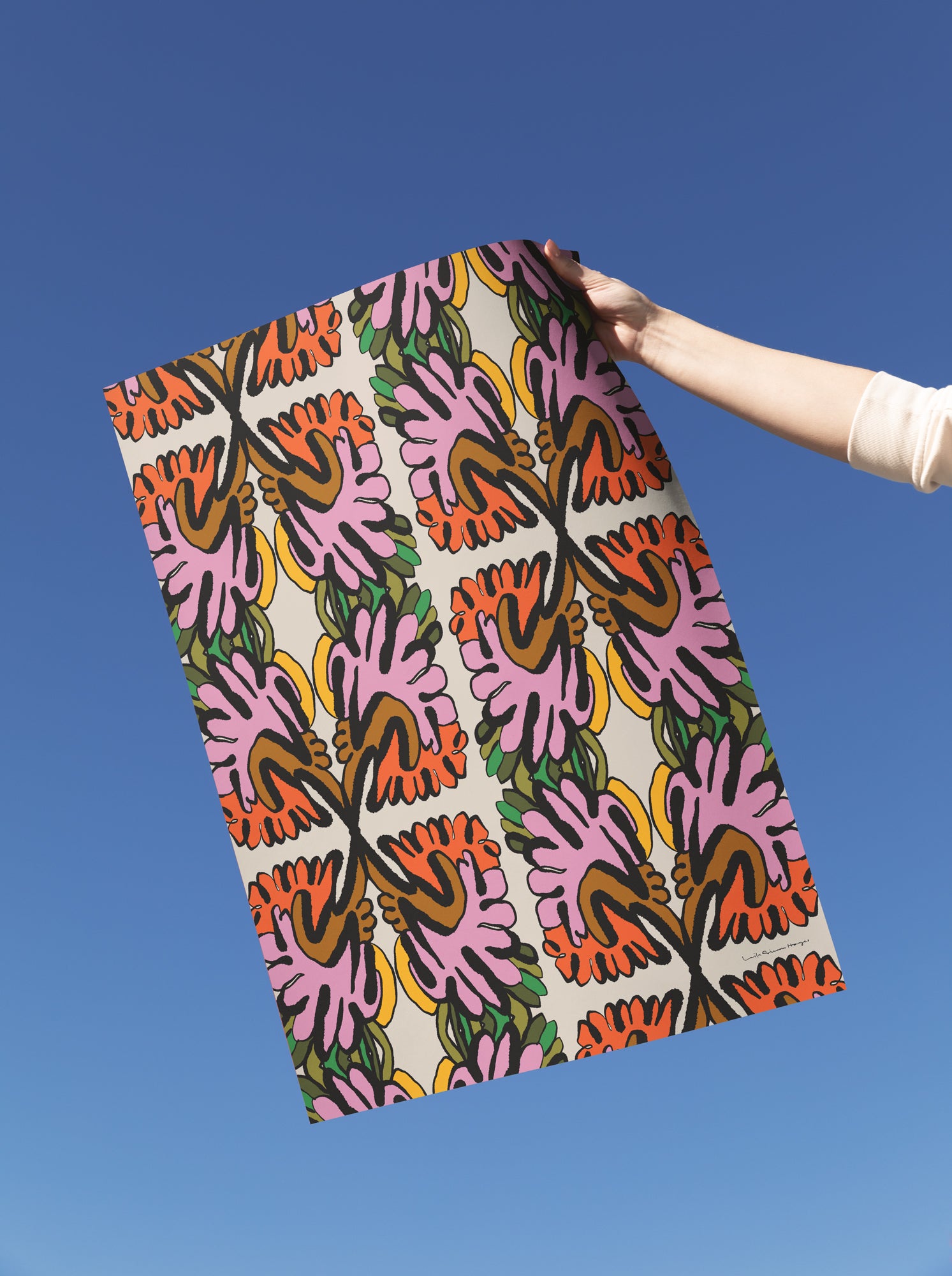 A person's arm extends into the frame holding fine art print against a clear blue sky. The paper features a vibrant, repeating ikat-style floral design in pink, orange, green, yellow, and black on a cream background. Designed by artist Leila Simon Hayes.