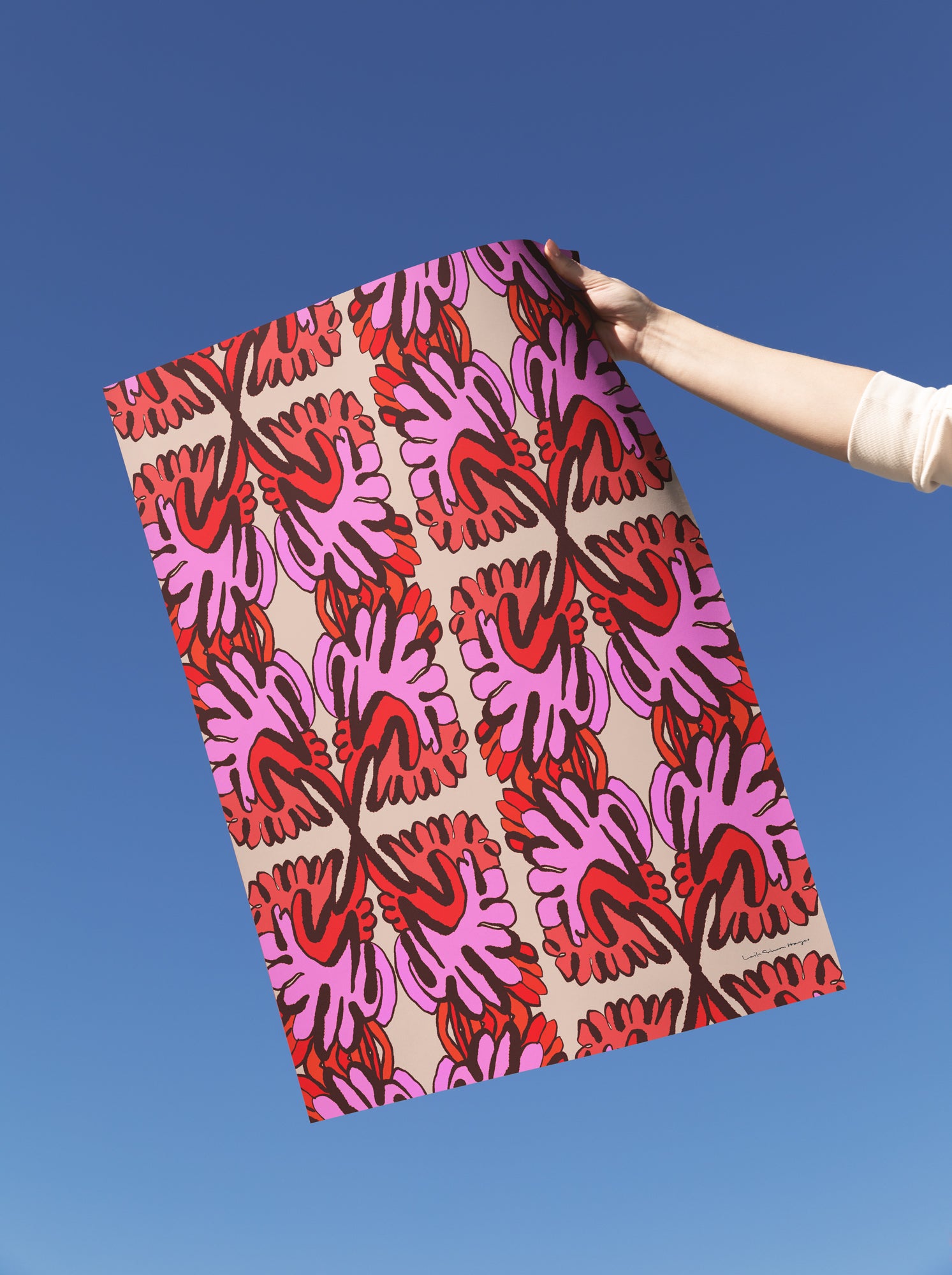 A hand holding up a fine art print with a bold retro ikat pattern in pink, red, orange, and beige against a clear blue sky. Designed by artist Leila Simon Hayes.