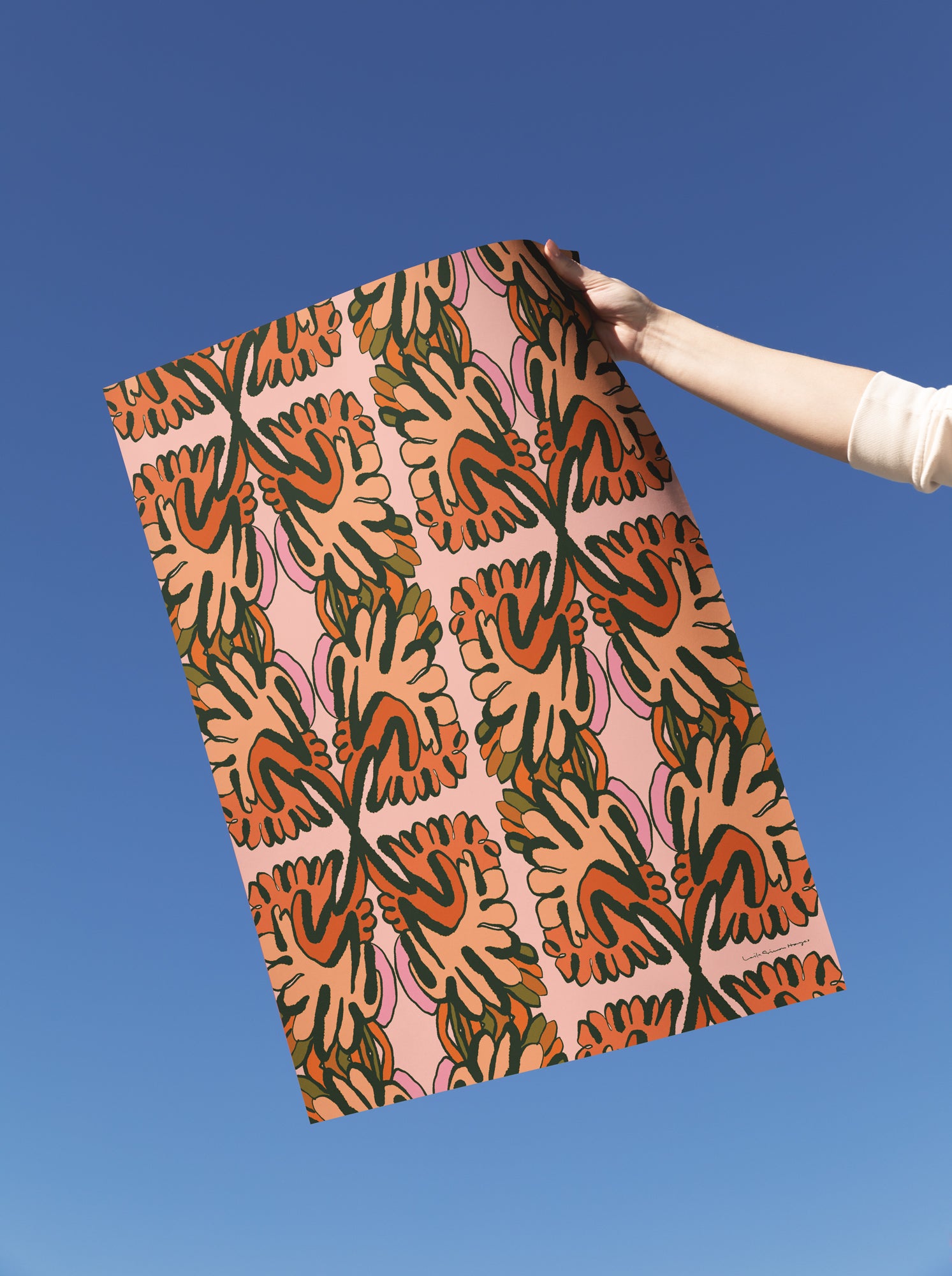 A person's arm holding up a fine art print against a clear blue sky. The pattern features retro, abstract paisley-like shapes in orange, dark green, and pink on a peach-beige background. Designed by artist Leila Simon Hayes.
