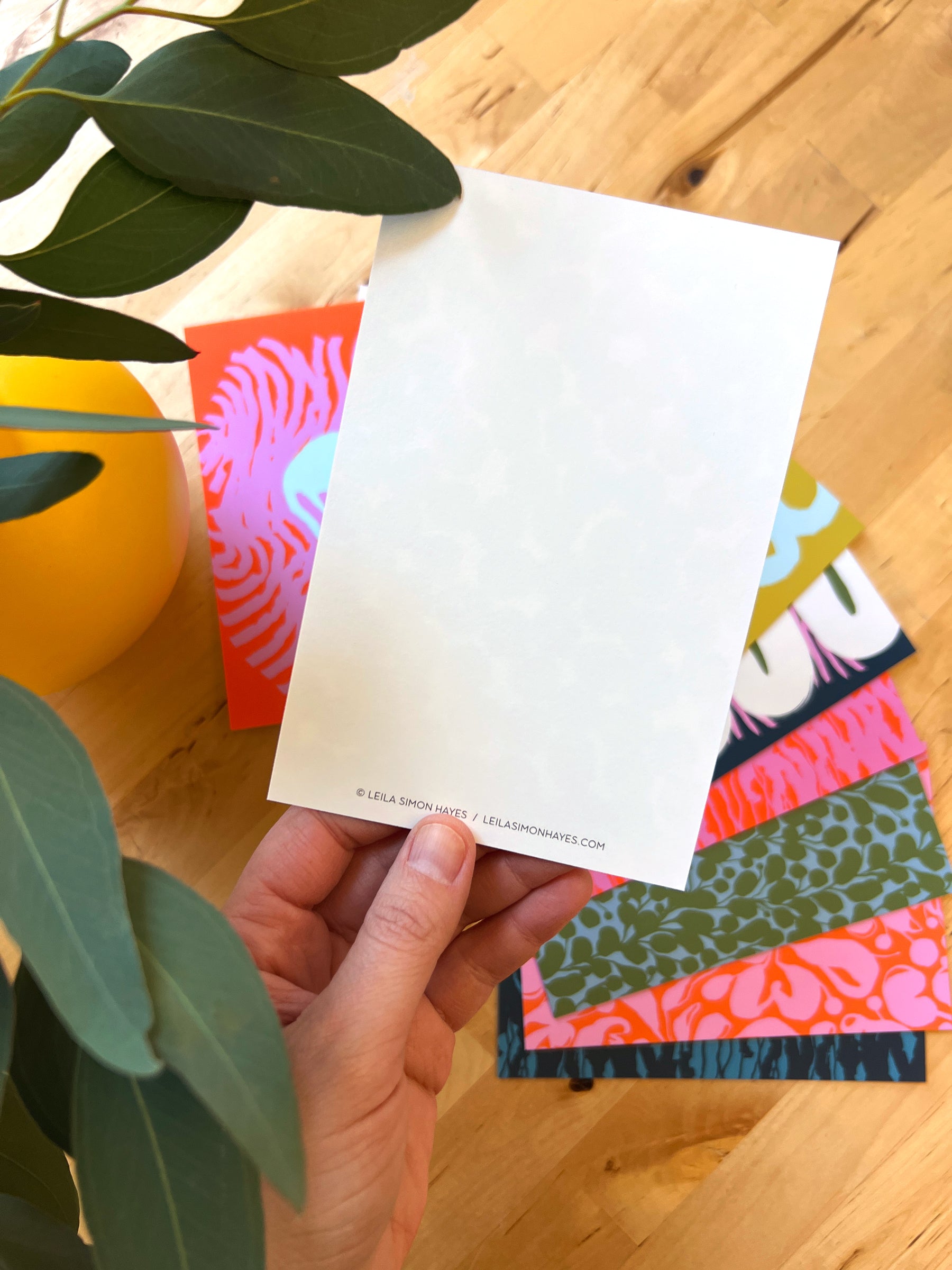 Person holding the back of a postcard to show the blank side. In the background there are 10 colorful and abstract postcards, designed by artist Leila Simon Hayes.