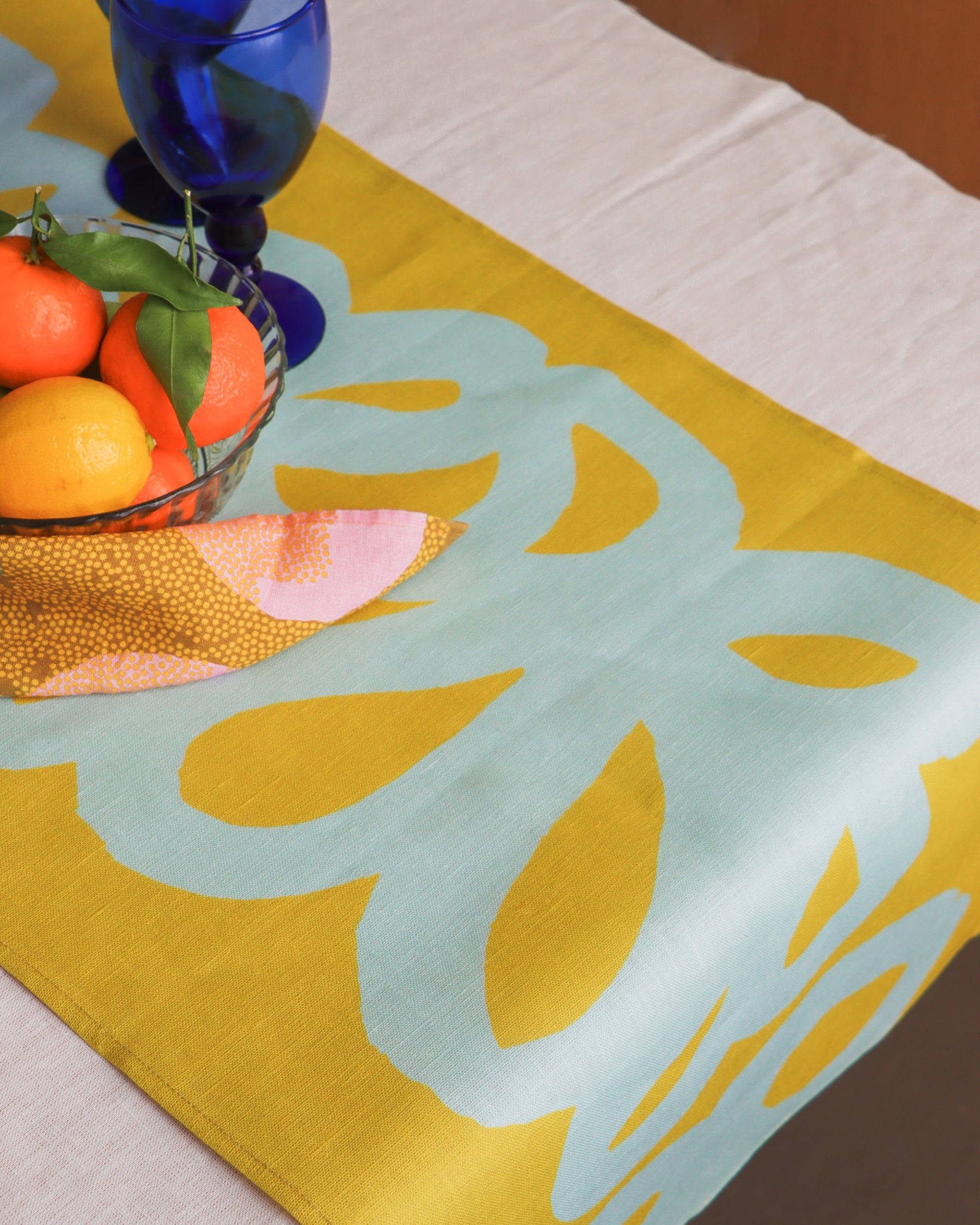 A green linen table runner with a floral loops pattern displayed on a table, part of a breakfast setup. Designed by artist Leila Simon Hayes.