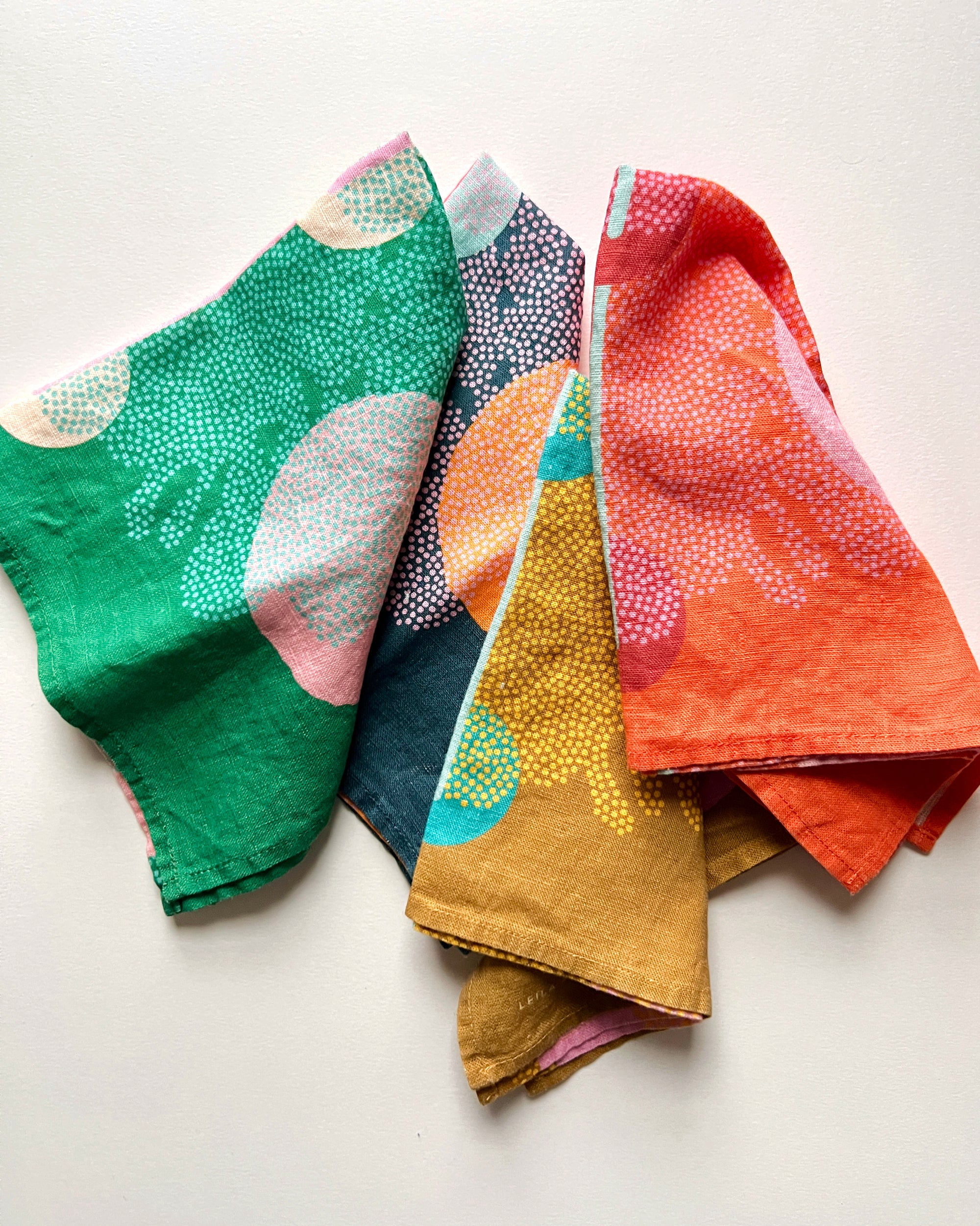 Four dinner napkins with the same abstract design in red, green, yellow and blue. Designed by artist Leila Simon Hayes, the napkins are folded and layered across a white background.