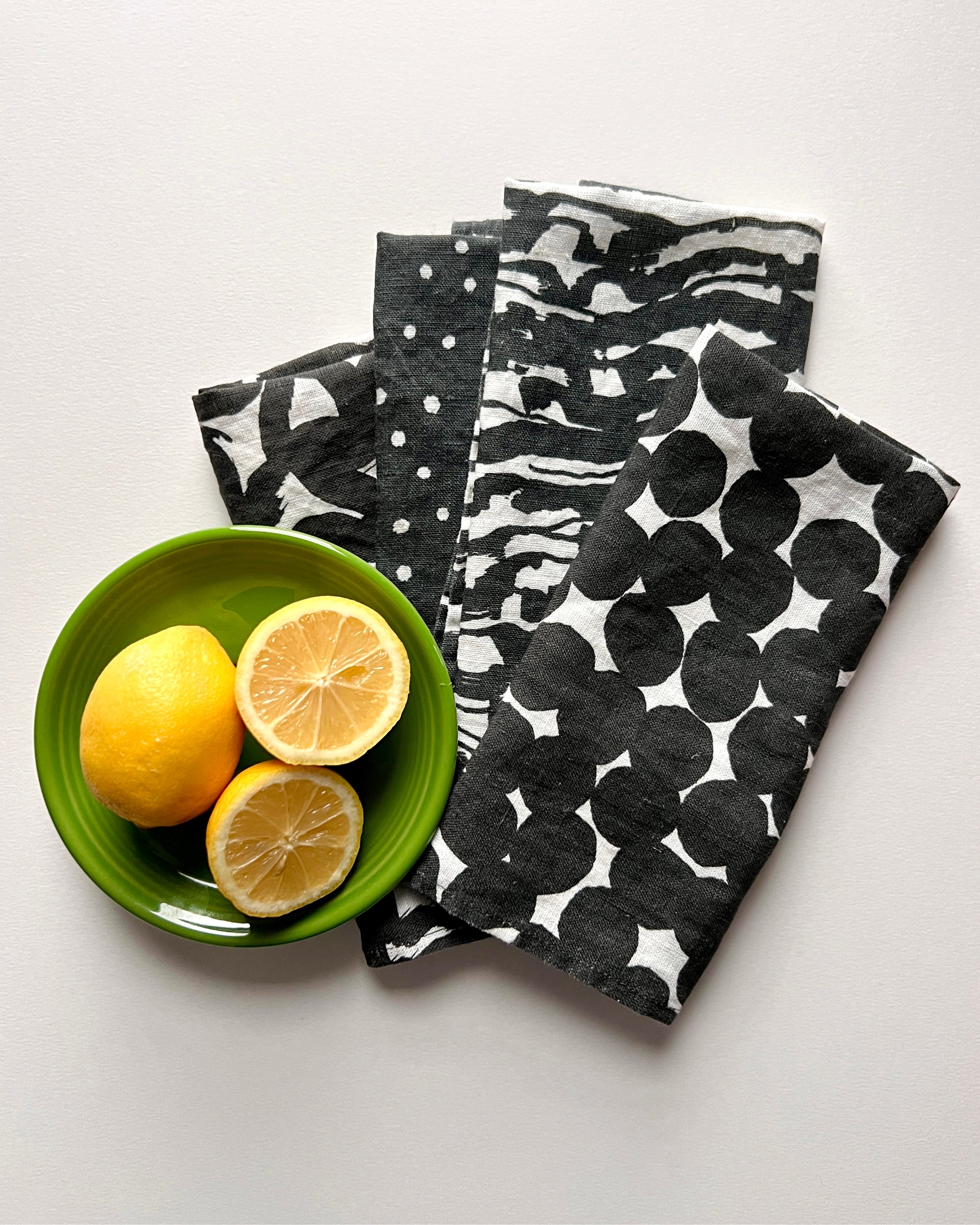 A set of four black and white linen napkins with a geometric pattern, displayed alongside a green bowl with two lemon halves. Designed by artist Leila Simon Hayes.