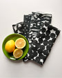 A set of four black and white linen napkins with a geometric pattern, displayed alongside a green bowl with two lemon halves. Designed by artist Leila Simon Hayes.