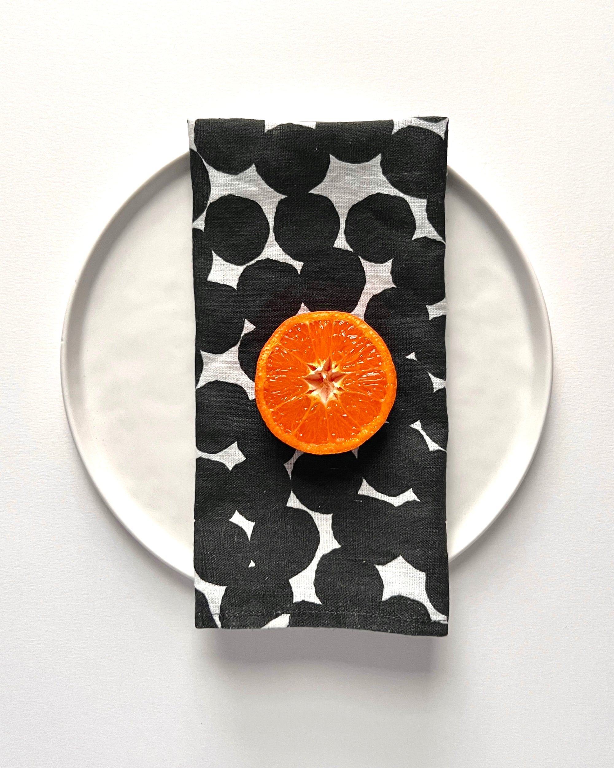 A white linen napkin with a dotted black pattern displayed on a white plate with half an orange on top. Designed by artist Leila Simon Hayes.