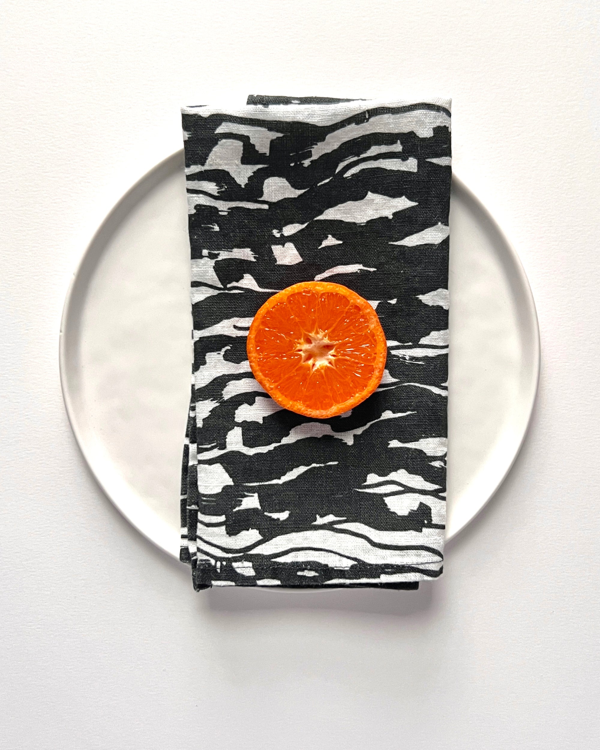 A black and white linen napkin with a wave pattern displayed on a white plate, with a half an orange placed on top for scale. Designed by artist Leila Simon Hayes.