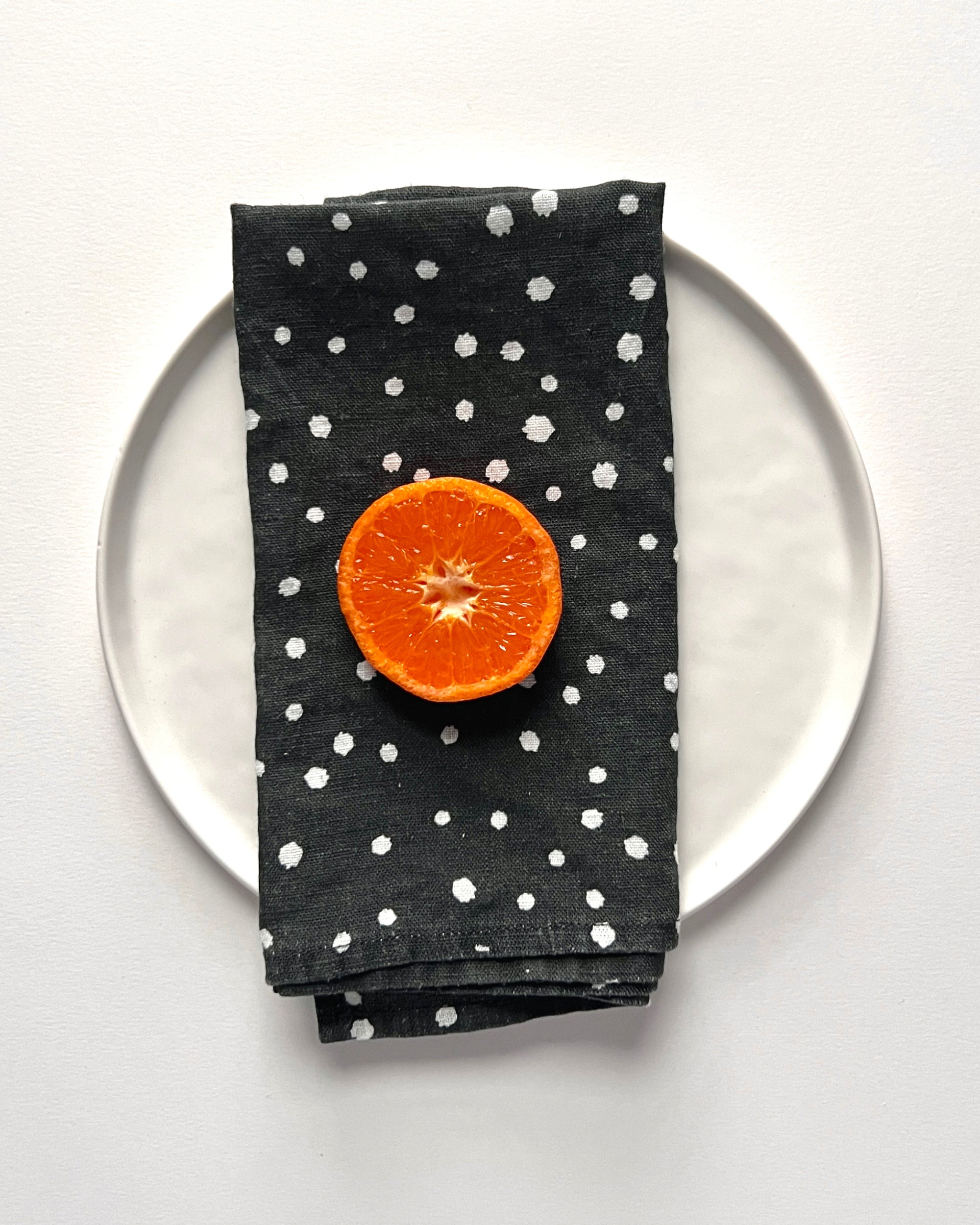 A black linen napkin with organic small white polka dots, displayed on a white plate with a half orange on top of it. Designed by artist Leila Simon Hayes