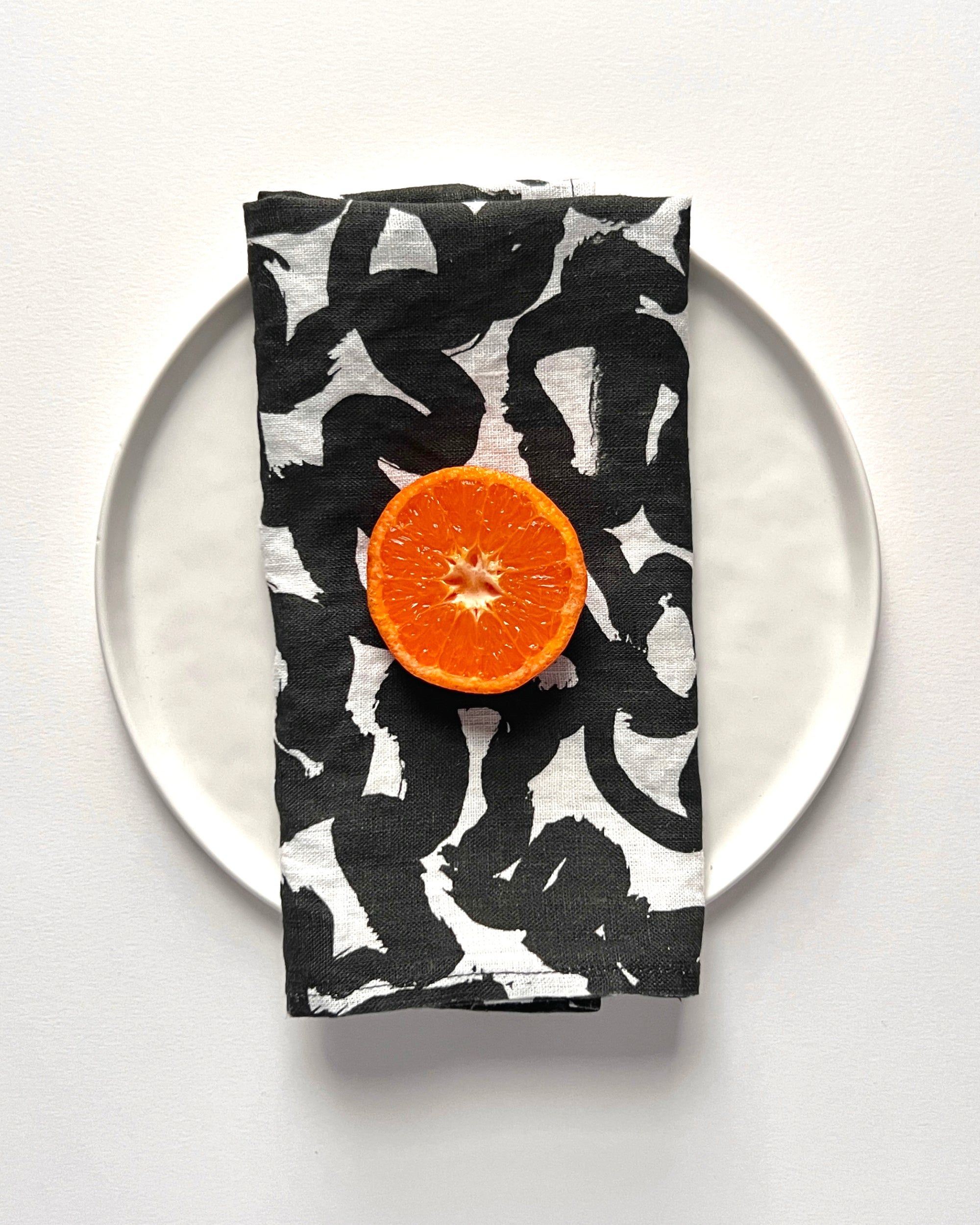 A white linen napkin with a black abstract paintbrush stroke pattern, designed by artist Leila Simon Hayes. Displayed on a white plate, with a slice of orange placed on top.
